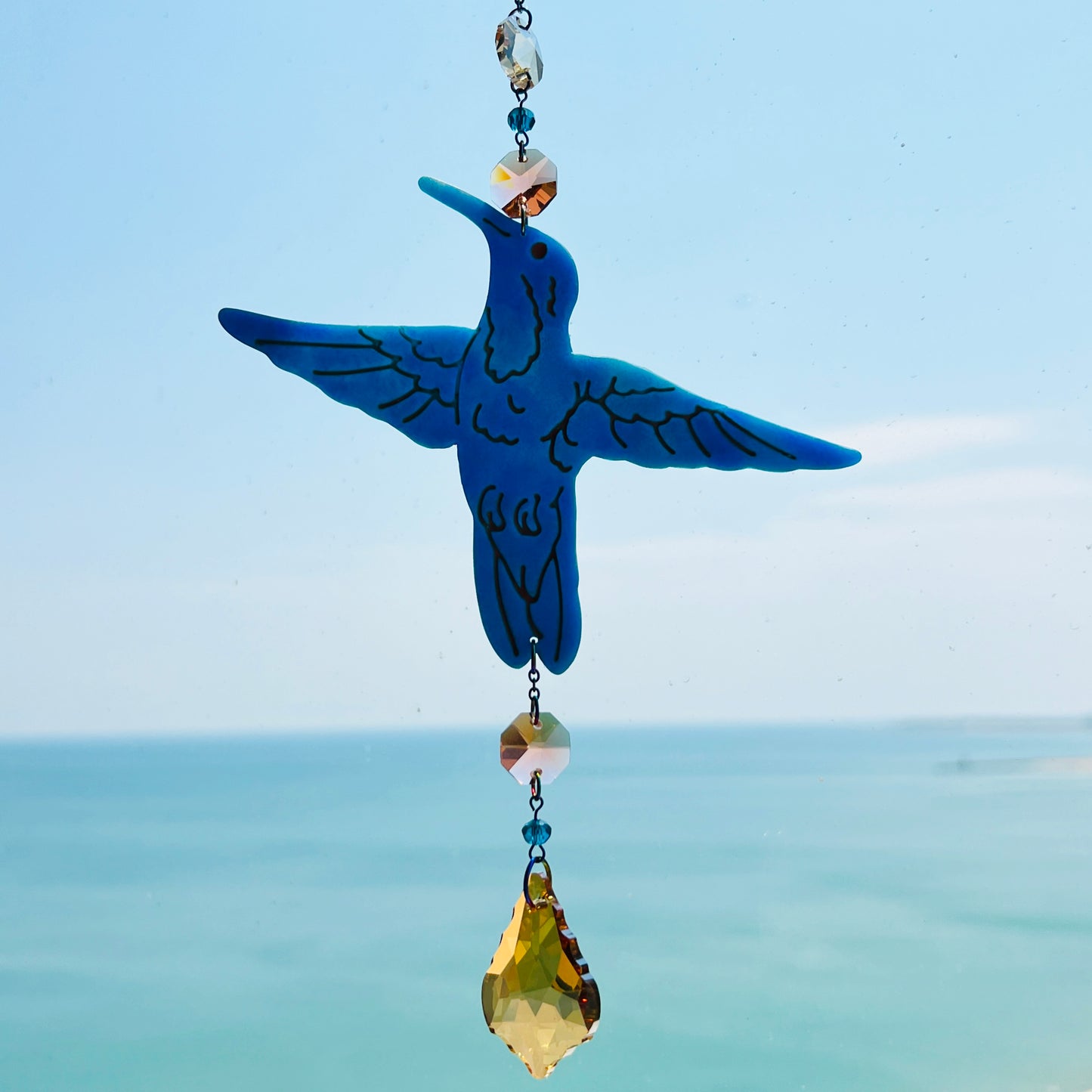 A handmade hummingbird suncatcher crafted from semi-transparent blue polymer clay, featuring a detailed hummingbird design suspended from a gold chain with a faceted champagne maple leaf glass crystal that casts rainbows in the sunlight. Perfect for nature lovers and whimsical window decor.