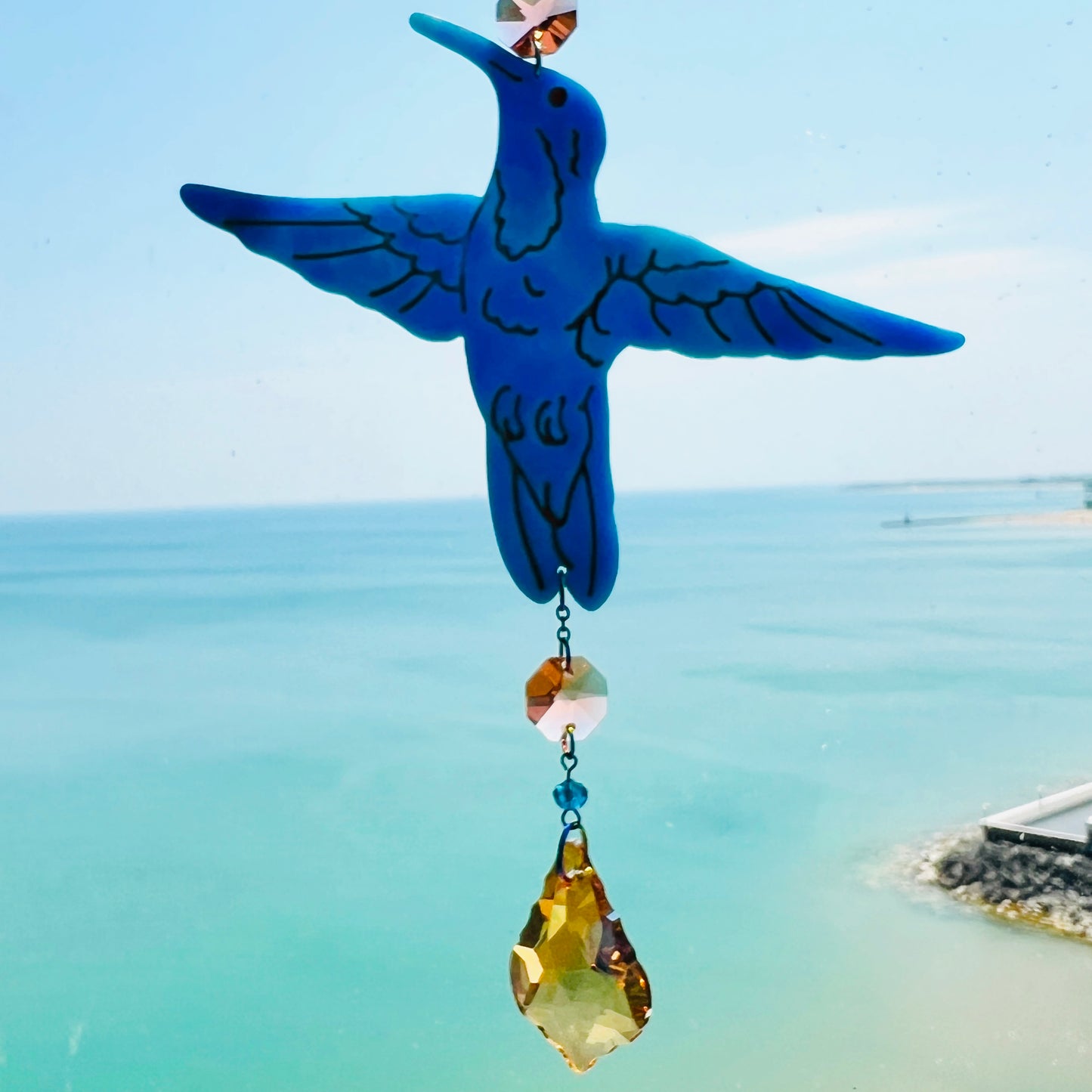 A handmade hummingbird suncatcher crafted from semi-transparent blue polymer clay, featuring a detailed hummingbird design suspended from a gold chain with a faceted champagne maple leaf glass crystal that casts rainbows in the sunlight. Perfect for nature lovers and whimsical window decor.