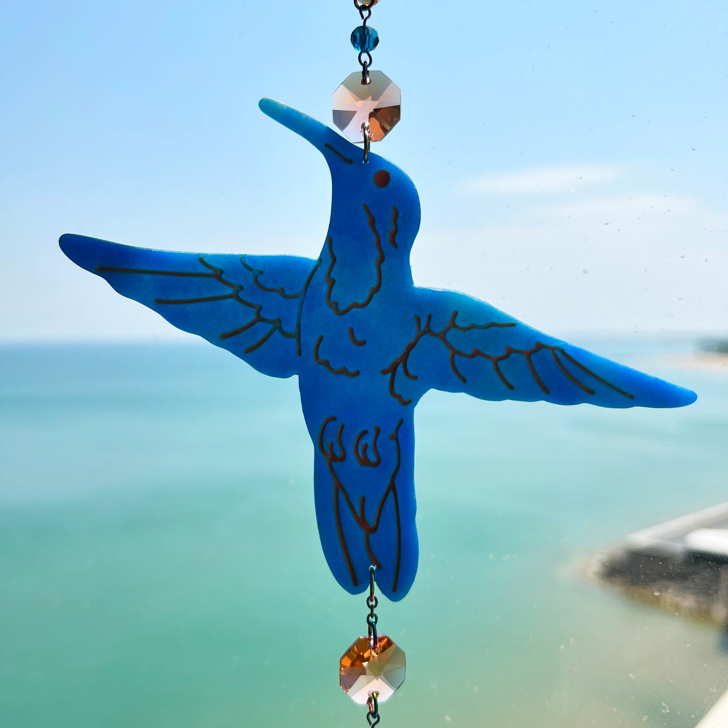 A handmade hummingbird suncatcher crafted from semi-transparent blue polymer clay, featuring a detailed hummingbird design suspended from a gold chain with a faceted champagne maple leaf glass crystal that casts rainbows in the sunlight. Perfect for nature lovers and whimsical window decor.