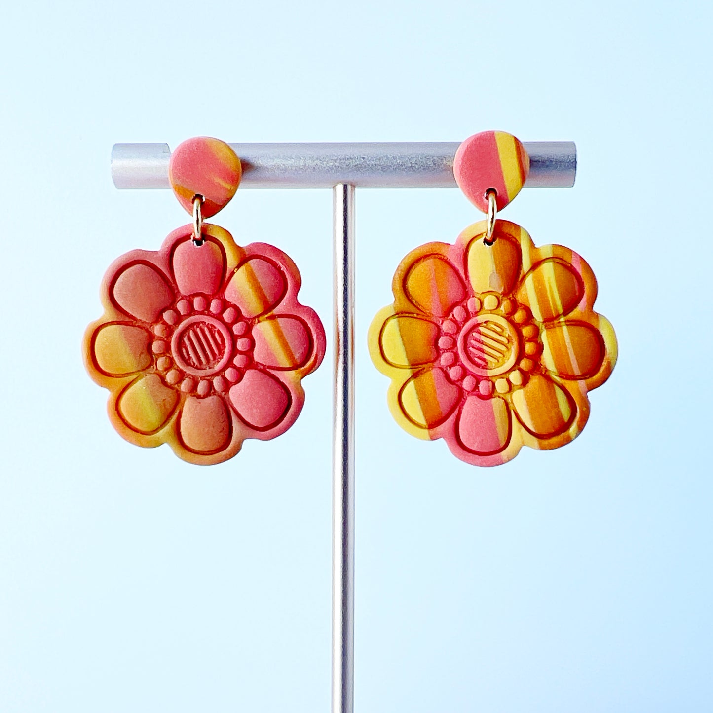 Handmade polymer clay earrings shaped like large, layered flowers with nine sculpted petals in a marbled blend of coral, orange, and yellow tones, finished with textured flower centers and coral clay stud tops on titanium posts