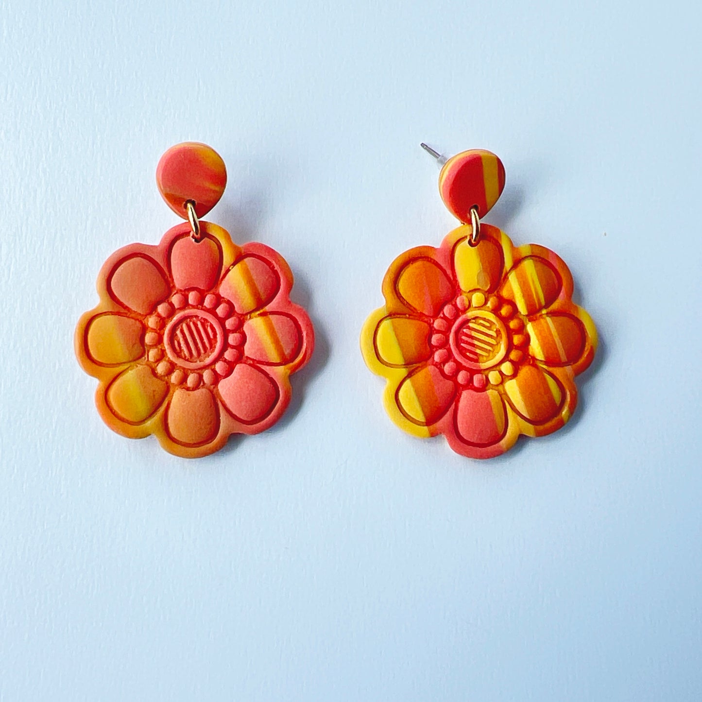Handmade polymer clay earrings shaped like large, layered flowers with nine sculpted petals in a marbled blend of coral, orange, and yellow tones, finished with textured flower centers and coral clay stud tops on titanium posts