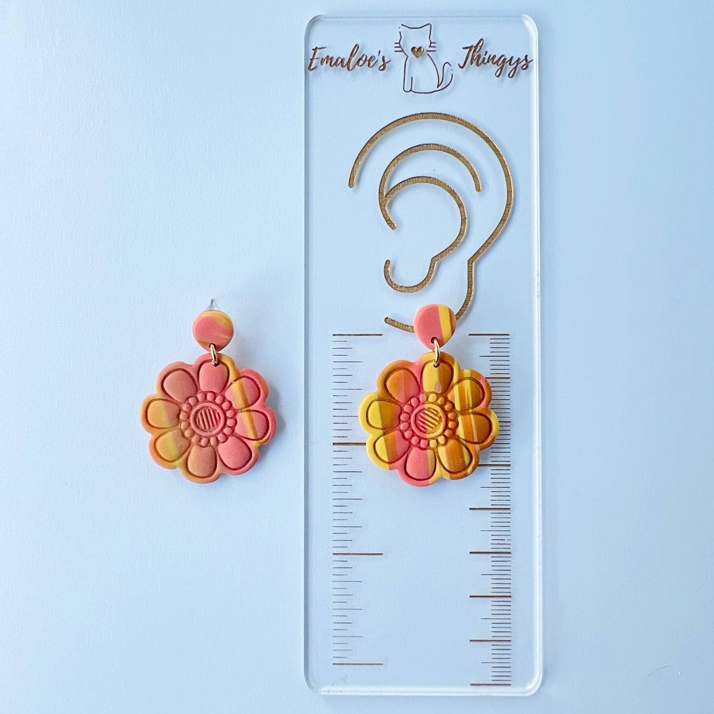 Handmade polymer clay earrings shaped like large, layered flowers with nine sculpted petals in a marbled blend of coral, orange, and yellow tones, finished with textured flower centers and coral clay stud tops on titanium posts. They are displayed on a branded ruler showing the length to be 1.75 inches long