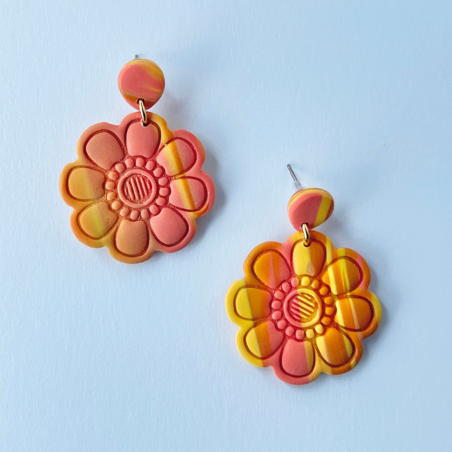 Handmade polymer clay earrings shaped like large, layered flowers with nine sculpted petals in a marbled blend of coral, orange, and yellow tones, finished with textured flower centers and coral clay stud tops on titanium posts