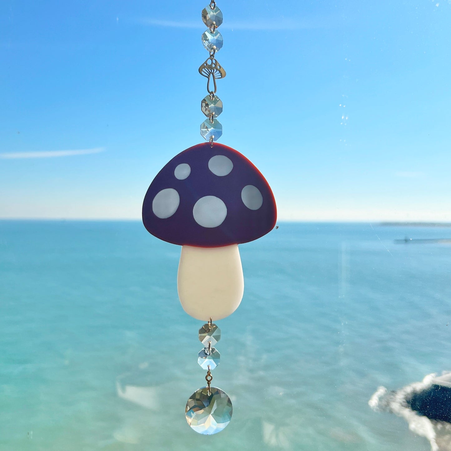 A whimsical handmade mushroom suncatcher crafted from polymer clay, featuring a brown and white spotted toadstool design suspended from a gold chain, with a dangling faceted glass crystal that casts rainbows in the sunlight. Ideal for woodland, fairycore, or cottagecore decor.