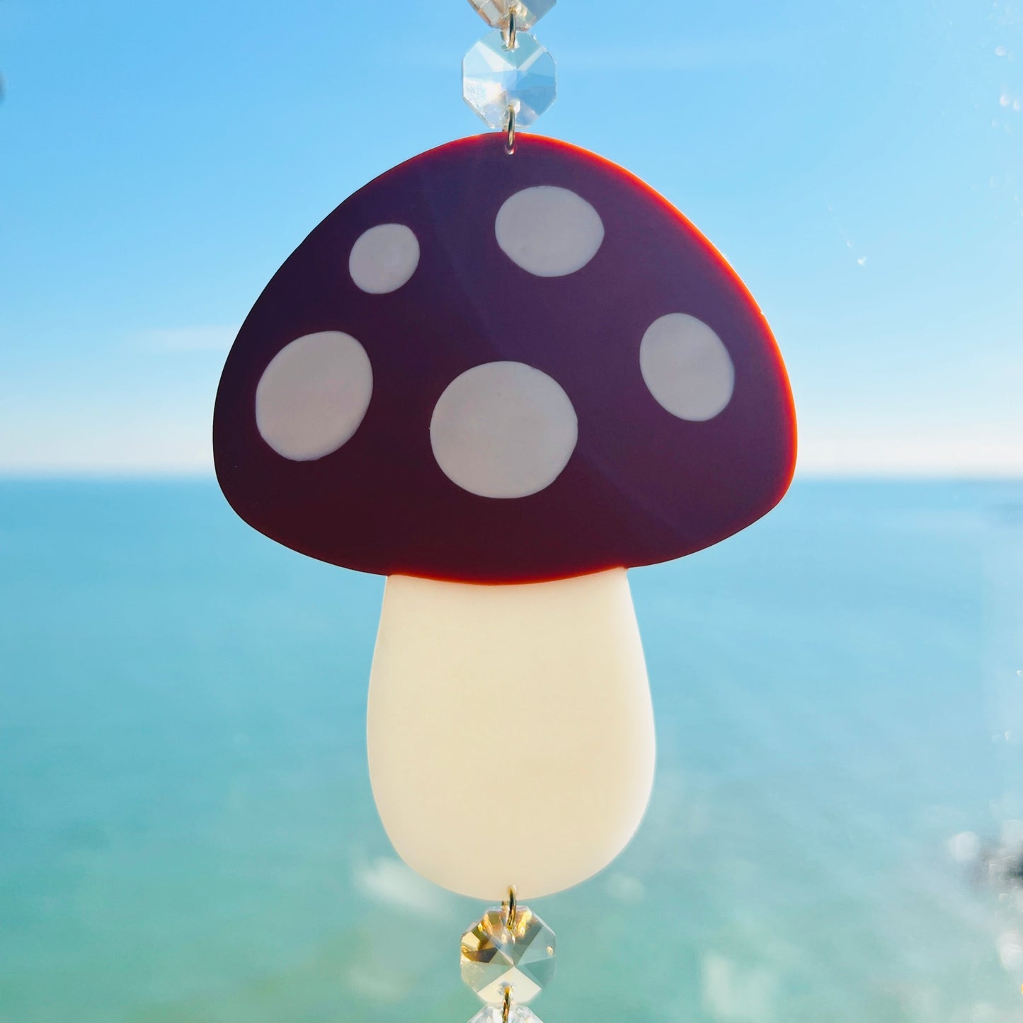 A whimsical handmade mushroom suncatcher crafted from polymer clay, featuring a brown and white spotted toadstool design suspended from a gold chain, with a dangling faceted glass crystal that casts rainbows in the sunlight. Ideal for woodland, fairycore, or cottagecore decor.
