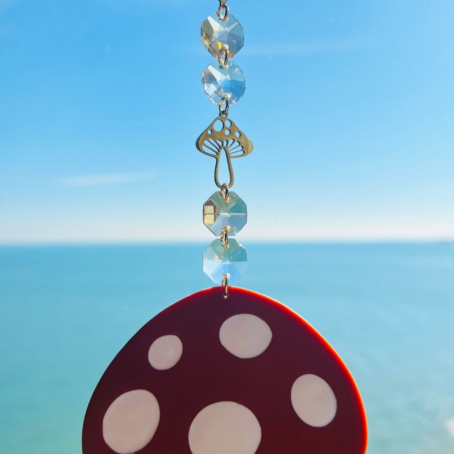 A whimsical handmade mushroom suncatcher crafted from polymer clay, featuring a brown and white spotted toadstool design suspended from a gold chain, with a dangling faceted glass crystal that casts rainbows in the sunlight. Ideal for woodland, fairycore, or cottagecore decor.