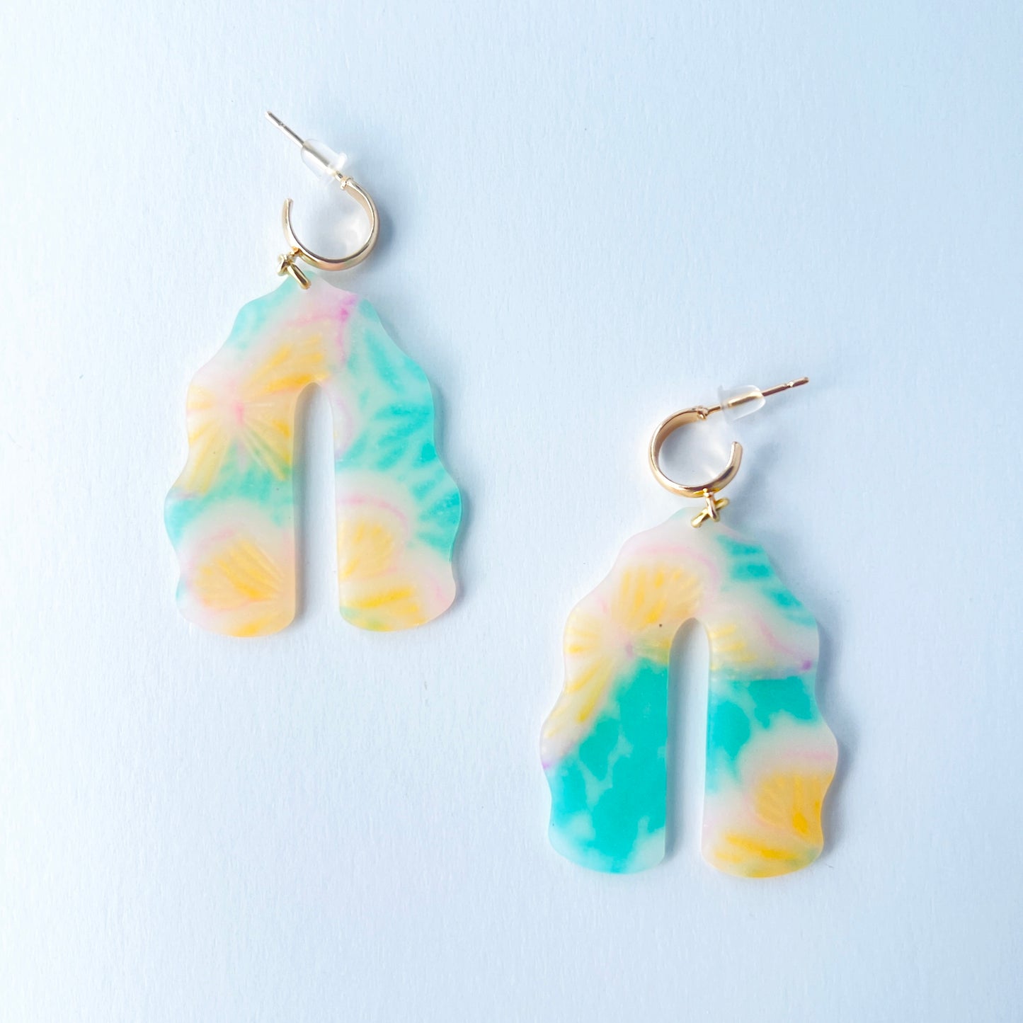 A pair of handmade polymer clay earrings shaped like arches, with swirled pastel colors and subtle details of yellow butterflies and green leafy accents. The semi-transparent clay design is paired with shiny gold-plated brass half hoop posts.