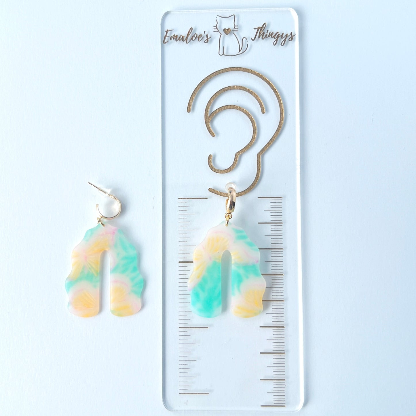 A pair of handmade polymer clay earrings shaped like arches, with swirled pastel colors and subtle details of yellow butterflies and green leafy accents. The semi-transparent clay design is paired with shiny gold-plated brass half hoop posts. One earring is laying on a clear branded ruler showing an approximate length of 2.25 inches