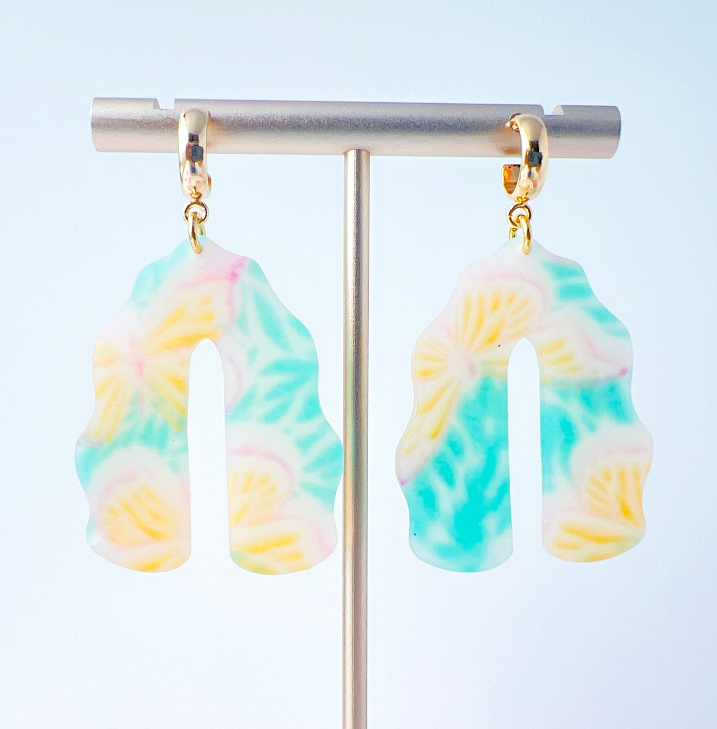 A pair of handmade polymer clay earrings shaped like arches, with swirled pastel colors and subtle details of yellow butterflies and green leafy accents. The semi-transparent clay design is paired with shiny gold-plated brass half hoop posts.