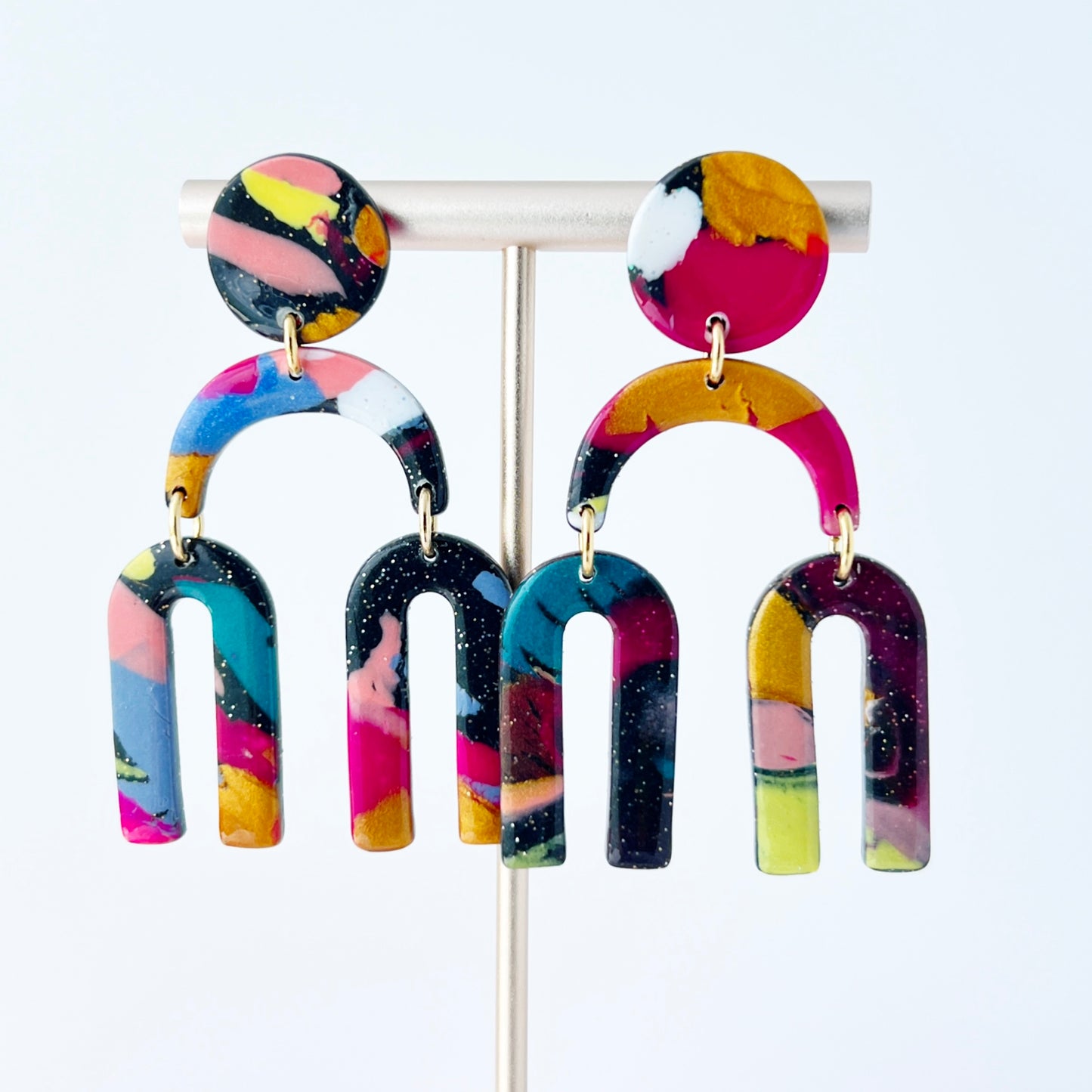 A vibrant pair of handmade polymer clay dangle earrings shaped like arched rainbows with U-shaped dangles. Marbled in a mix of pink, teal, gold, white, black, and glitter, with titanium posts. Displayed against a white background