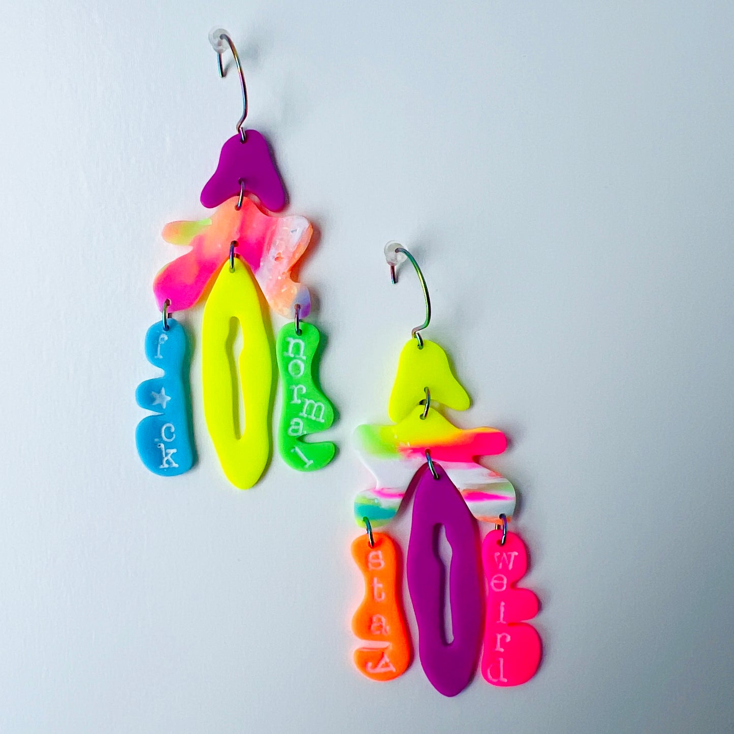 A pair of mismatched handmade statement earrings made from brightly colored neon polymer clay in layered abstract shapes. The left earring includes shapes in purple, orange, yellow, blue, and green and spells “F*ck Normal.” The right earring features neon pink, yellow, orange, purple, and green elements spelling “Stay Weird.” Both earrings are suspended from rainbow-plated stainless steel earwires and shown on a flat white background