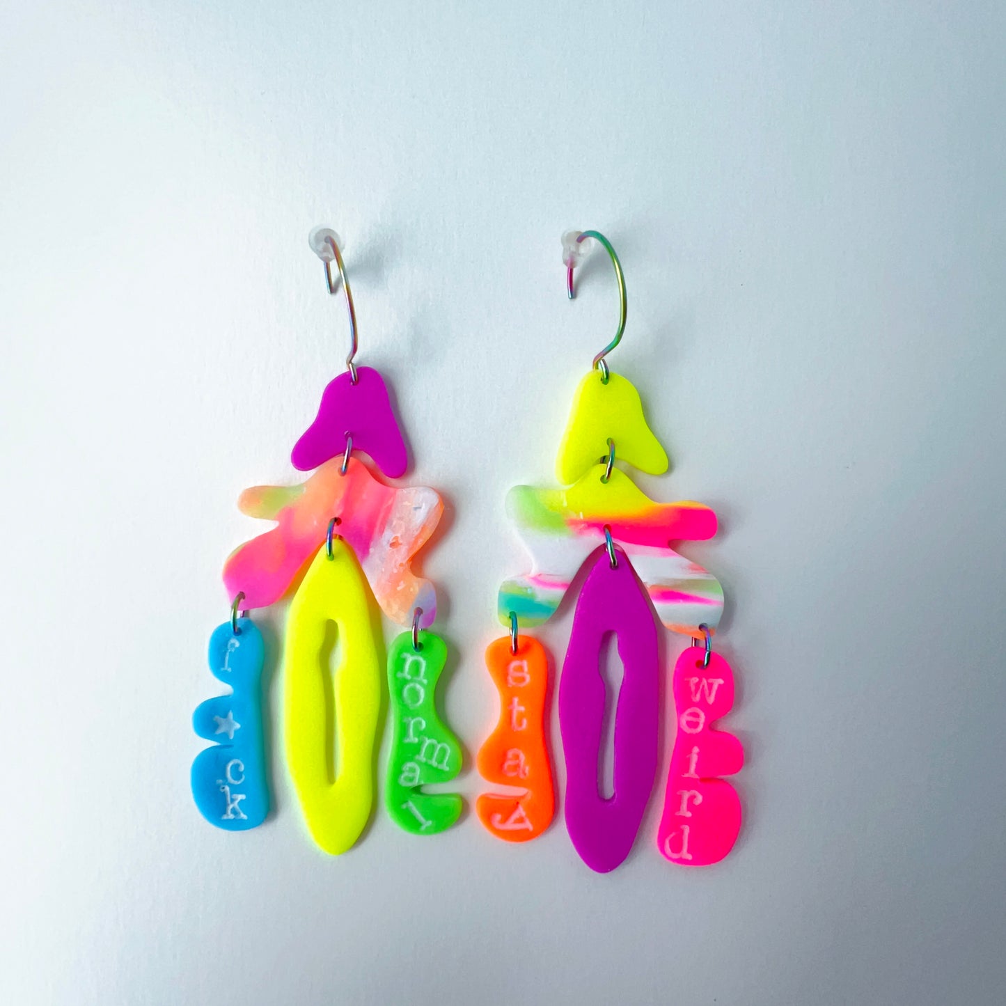 A pair of mismatched handmade statement earrings made from brightly colored neon polymer clay in layered abstract shapes. The left earring includes shapes in purple, orange, yellow, blue, and green and spells “F*ck Normal.” The right earring features neon pink, yellow, orange, purple, and green elements spelling “Stay Weird.” Both earrings are suspended from rainbow-plated stainless steel earwires and shown on a flat white background
