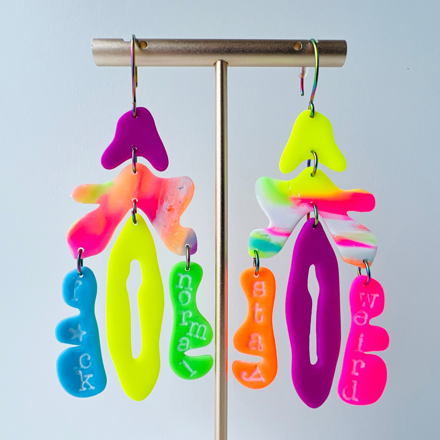 A pair of mismatched handmade statement earrings made from brightly colored neon polymer clay in layered abstract shapes hanging from a gold T-shaped bar. The left earring includes shapes in purple, orange, yellow, blue, and green and spells “F*ck Normal.” The right earring features neon pink, yellow, orange, purple, and green elements spelling “Stay Weird.” Both earrings are suspended from rainbow-plated stainless steel earwires and shown on a flat white backgroud