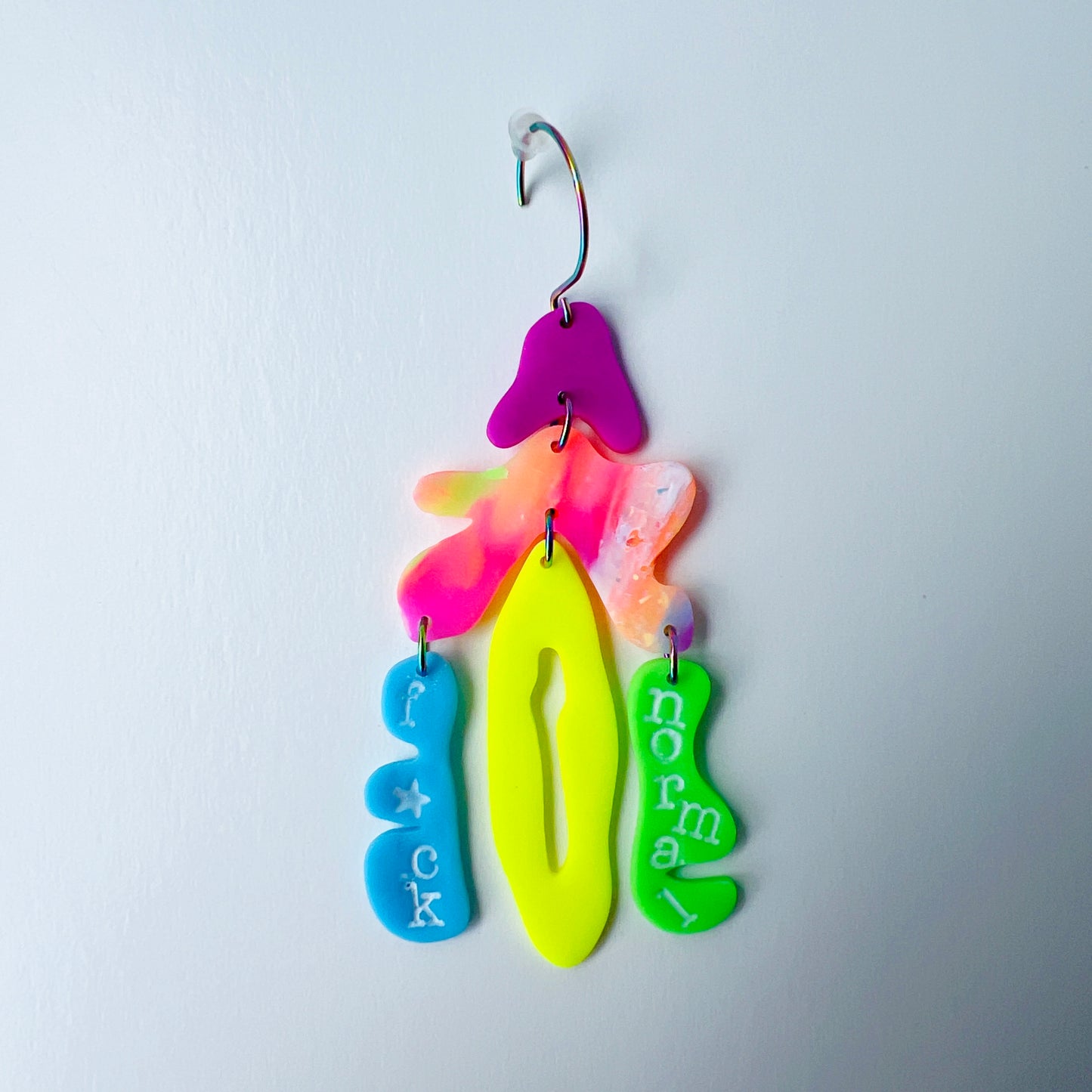 Left side from a mismatched handmade statement earrings that are made from brightly colored neon polymer clay in layered abstract shapes. The left earring includes shapes in purple, orange, yellow, blue, and green and spells “F*ck Normal.” Both earrings are suspended from rainbow-plated stainless steel earwires and shown on a flat white background
