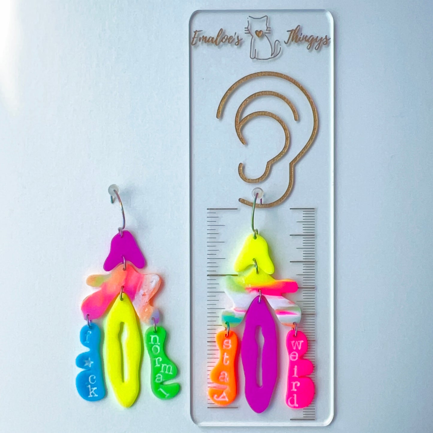 A pair of mismatched handmade statement earrings made from brightly colored neon polymer clay in layered abstract shapes. The left earring includes shapes in purple, orange, yellow, blue, and green and spells “F*ck Normal.” The right earring features neon pink, yellow, orange, purple, and green elements spelling “Stay Weird.” Both earrings are suspended from rainbow-plated stainless steel earwires and shown on a flat white background and with a a ruler, showing the approximate length of 3.5 inches/88.9 mm