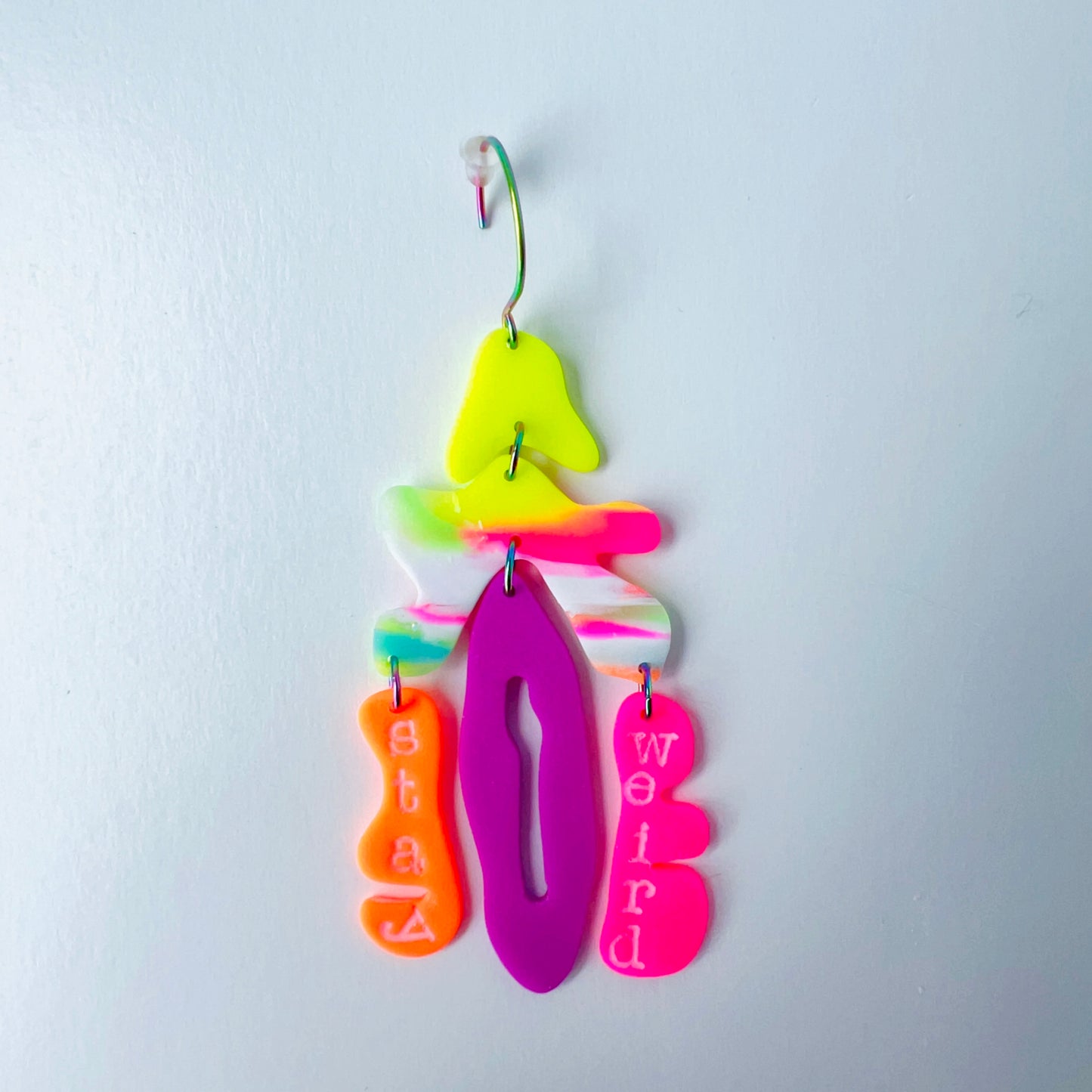 Right side from a mismatched handmade statement earrings that are made from brightly colored neon polymer clay in layered abstract shapes. The right earring features neon pink, yellow, orange, purple, and green elements spelling “Stay Weird.” Both earrings are suspended from rainbow-plated stainless steel earwires and shown on a flat white background
