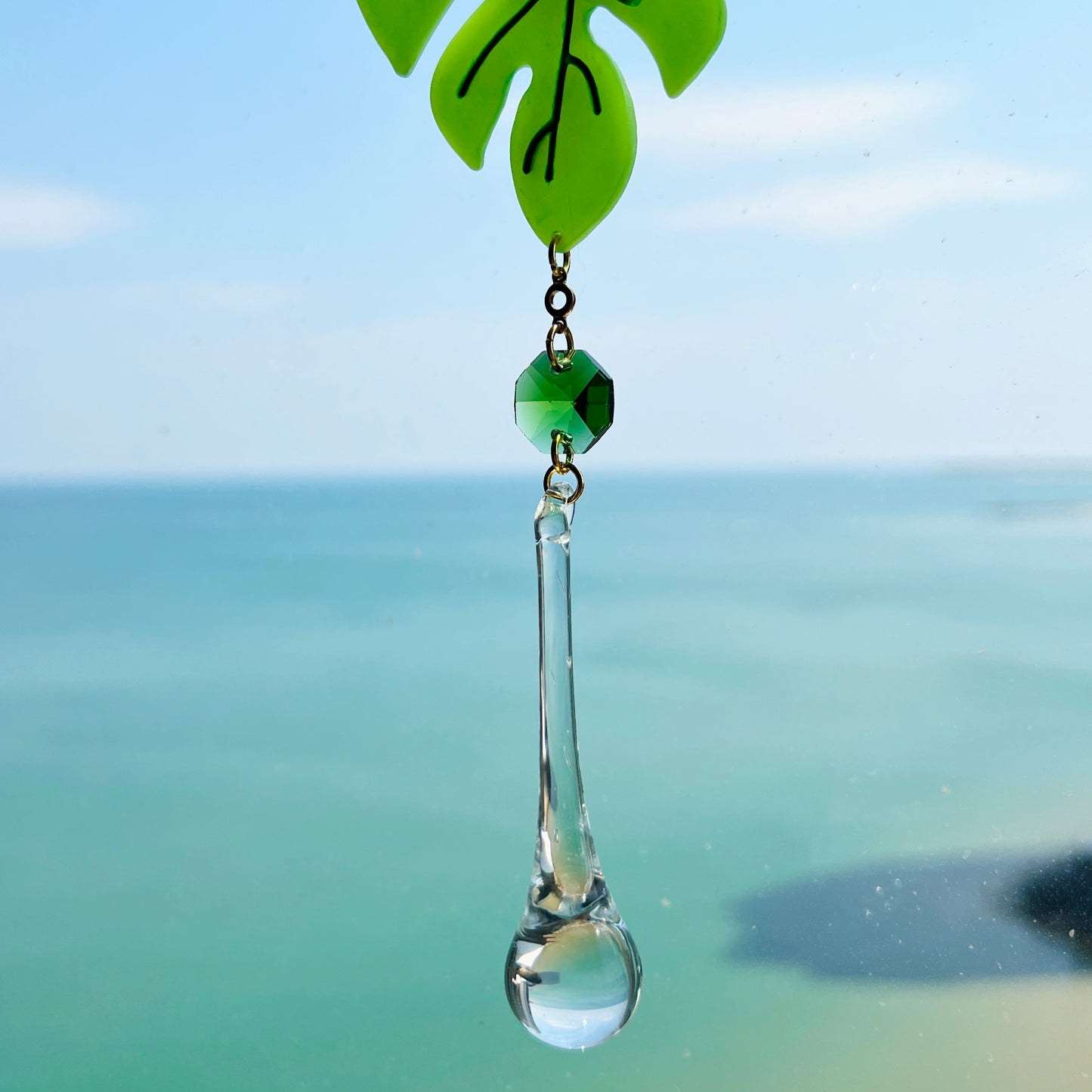 A handmade monstera leaf suncatcher made from green polymer clay with detailed vein textures, hanging from a gold loop and chain, featuring a faceted glass crystal that casts rainbows in sunlight. Ideal for botanical or boho home decor.