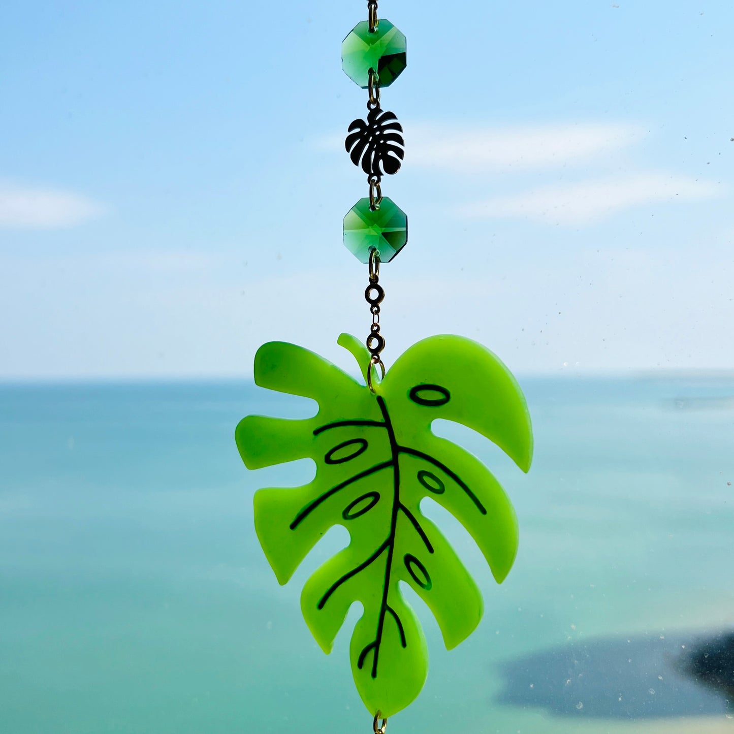 A handmade monstera leaf suncatcher made from green polymer clay with detailed vein textures, hanging from a gold loop and chain, featuring a faceted glass crystal that casts rainbows in sunlight. Ideal for botanical or boho home decor.