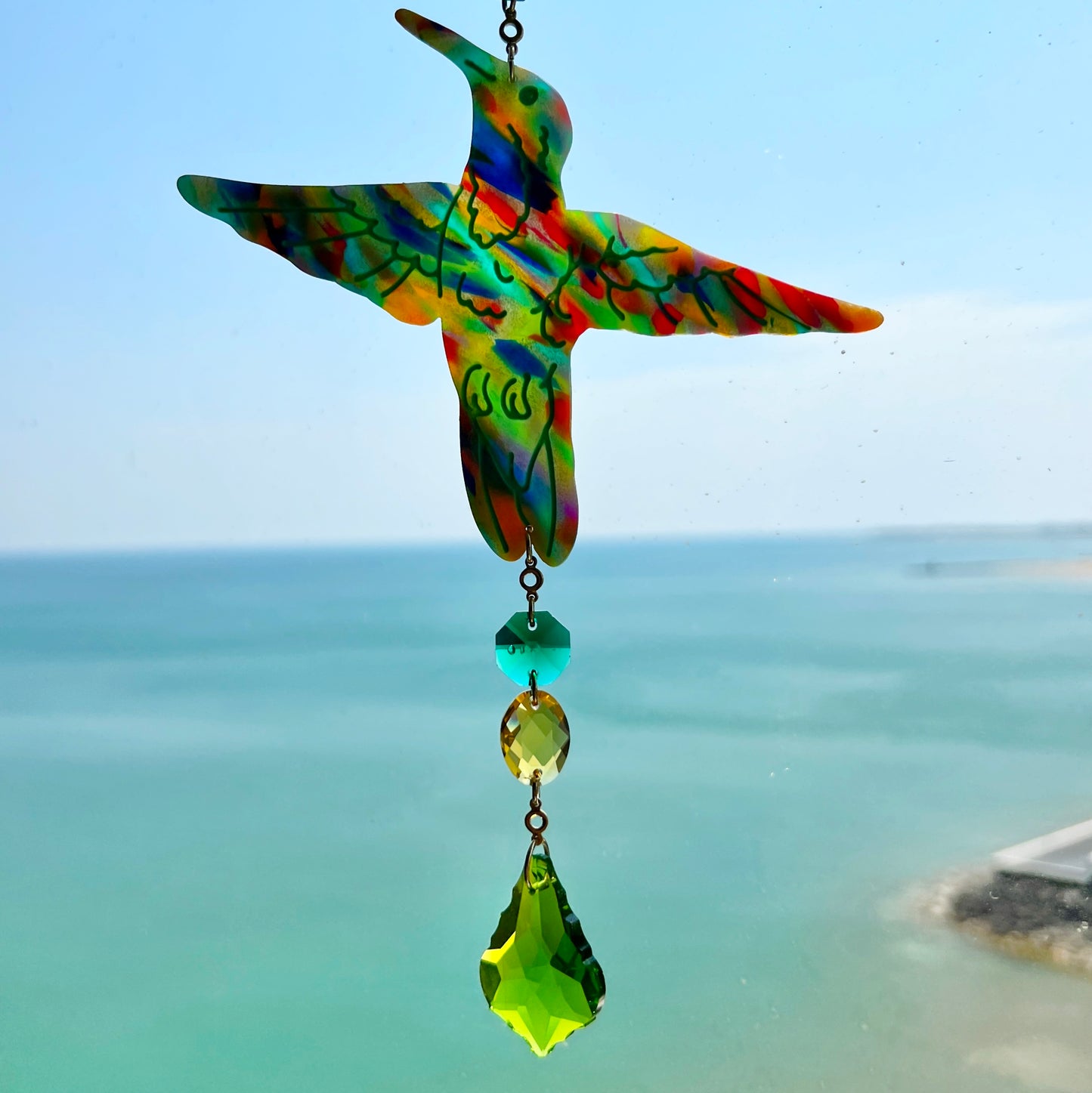 A handmade hummingbird suncatcher crafted from colorful polymer clay, featuring a detailed hummingbird design in shades of green, blue, and pink, suspended from a gold chain with a faceted green maple leaf glass crystal that casts rainbows in the sunlight. Perfect for nature lovers and whimsical window decor.