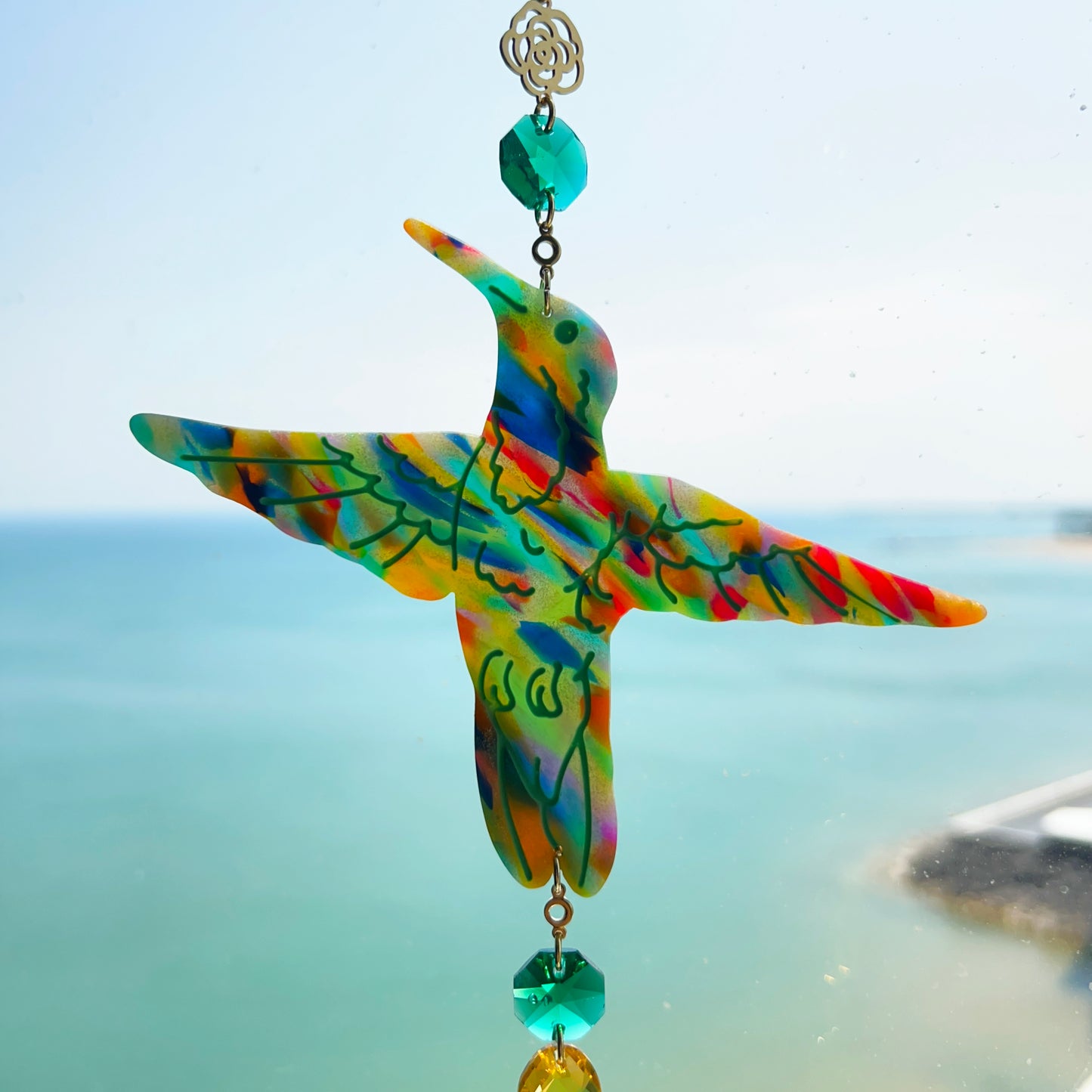 A handmade hummingbird suncatcher crafted from colorful polymer clay, featuring a detailed hummingbird design in shades of green, blue, and pink, suspended from a gold chain with a faceted green maple leaf glass crystal that casts rainbows in the sunlight. Perfect for nature lovers and whimsical window decor.