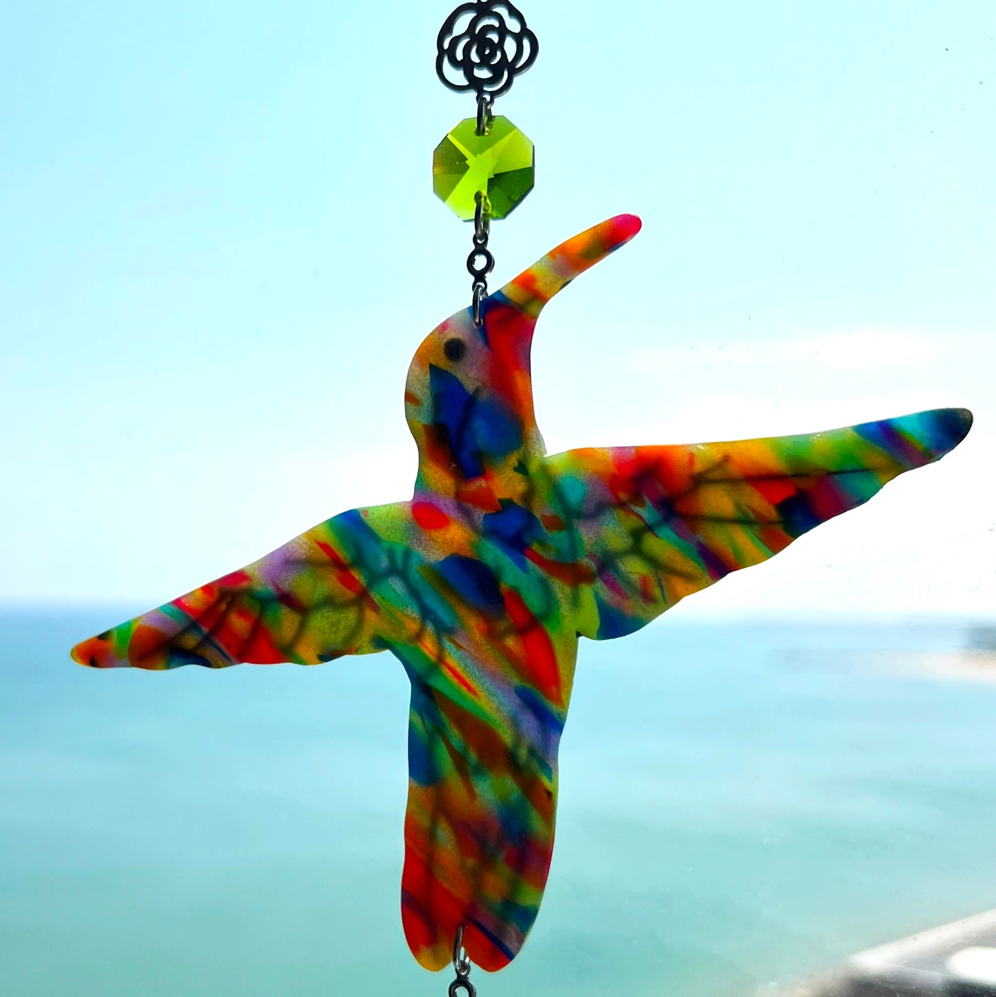 A handmade hummingbird suncatcher crafted from colorful polymer clay, featuring a detailed hummingbird design in shades of green, blue, and pink, suspended from a gold chain with a faceted blue maple leaf glass crystal that casts rainbows in the sunlight. Perfect for nature lovers and whimsical window decor.