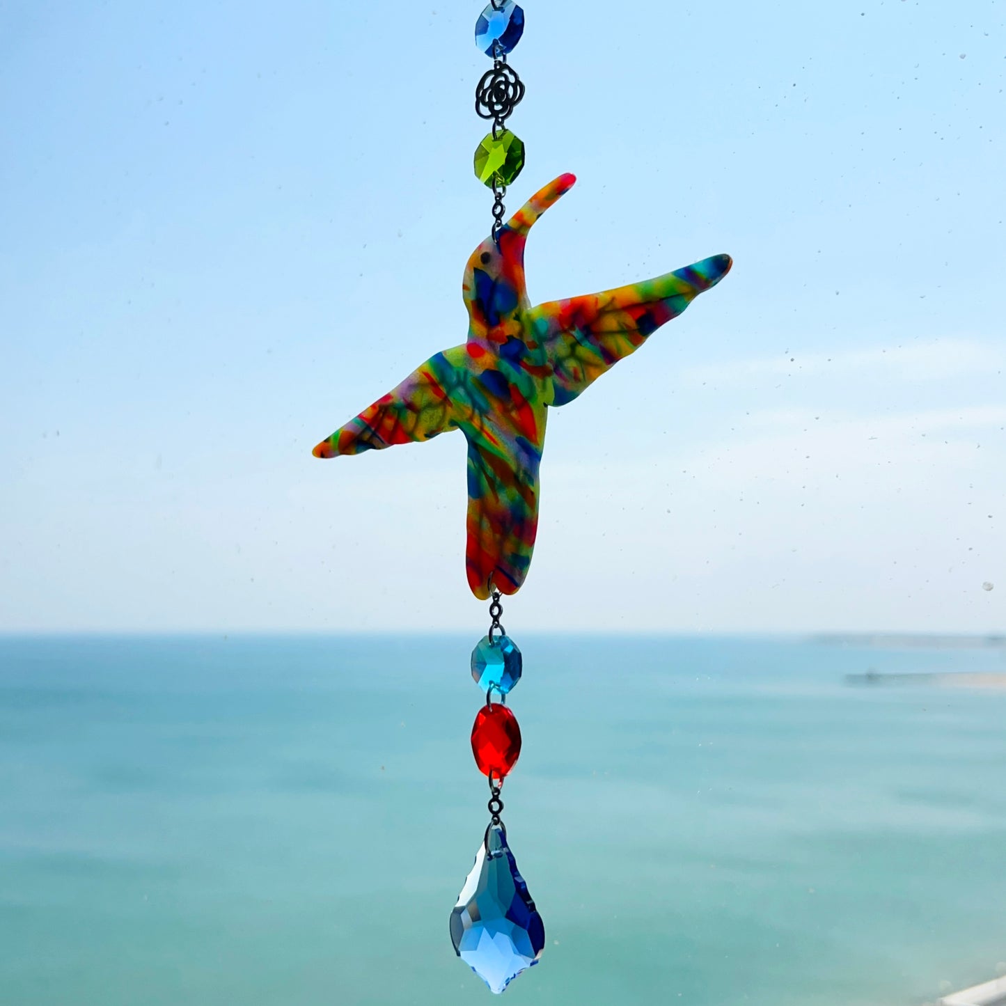 A handmade hummingbird suncatcher crafted from colorful polymer clay, featuring a detailed hummingbird design in shades of green, blue, and pink, suspended from a gold chain with a faceted blue maple leaf glass crystal that casts rainbows in the sunlight. Perfect for nature lovers and whimsical window decor.