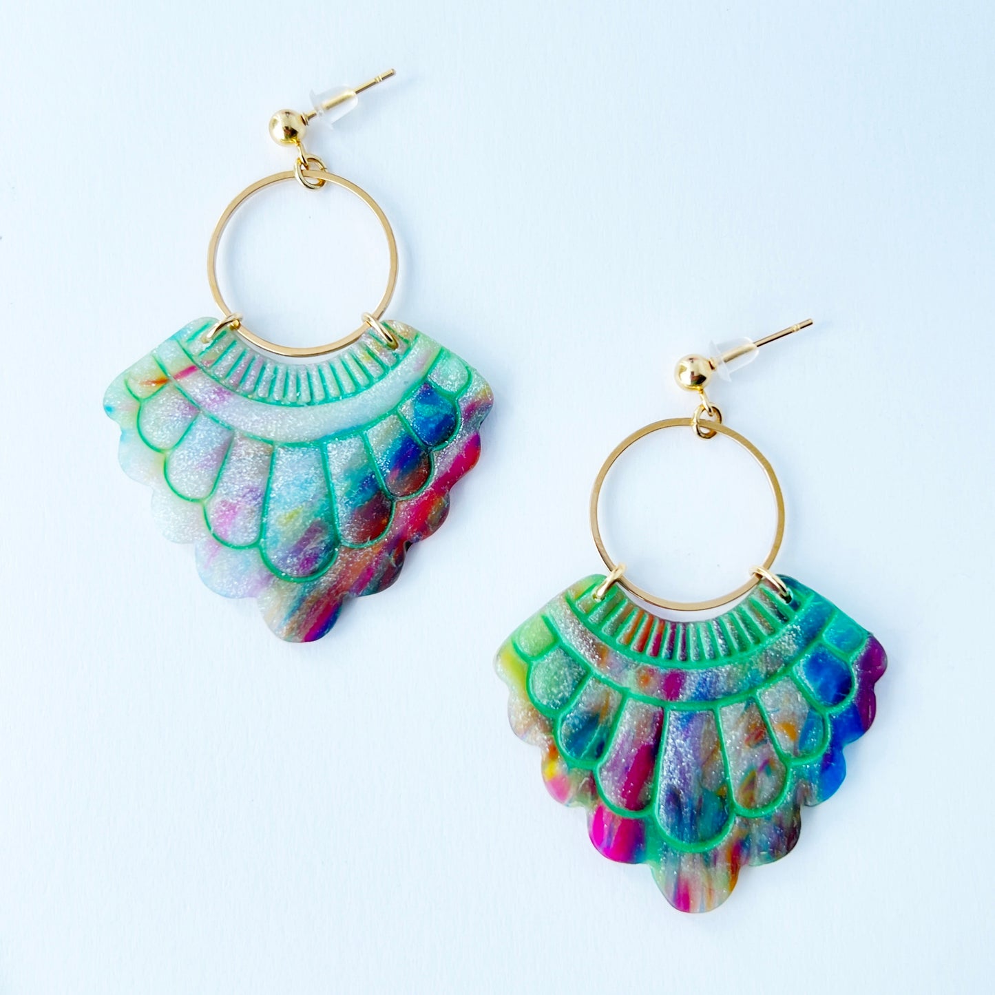 A pair of handmade fan-shaped polymer clay earrings displayed on a white background. The earrings feature gold-plated ball posts and hoops, with semi-transparent rainbow-colored clay and green etched fan-like lines. The marbling effect shows swirls of pink, blue, green, orange, and white.