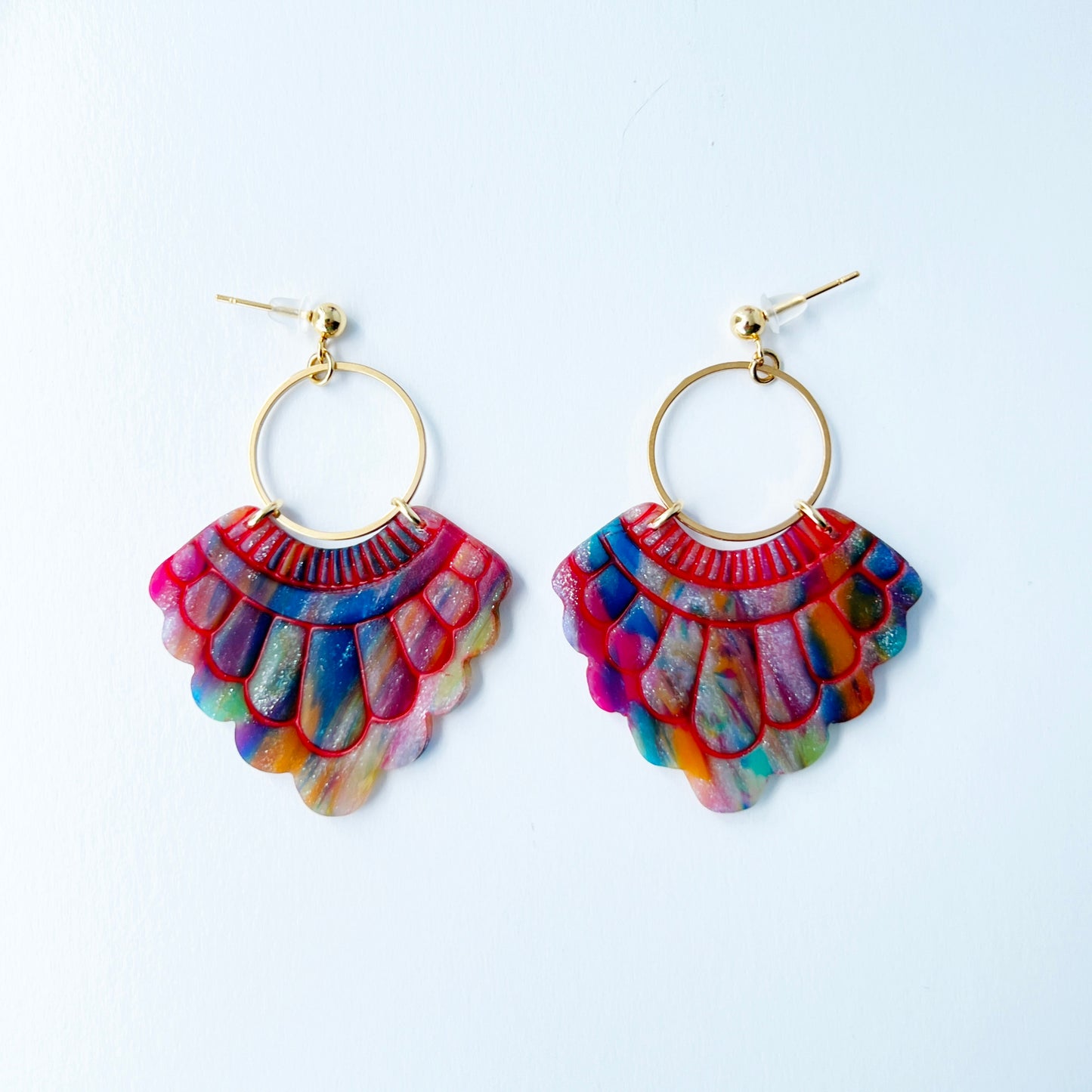 A pair of handmade fan-shaped polymer clay earrings displayed on a white background. The earrings feature gold-plated ball posts and hoops, with semi-transparent rainbow-colored clay and red etched fan-like lines. The marbling effect shows swirls of pink, blue, green, orange, and white.