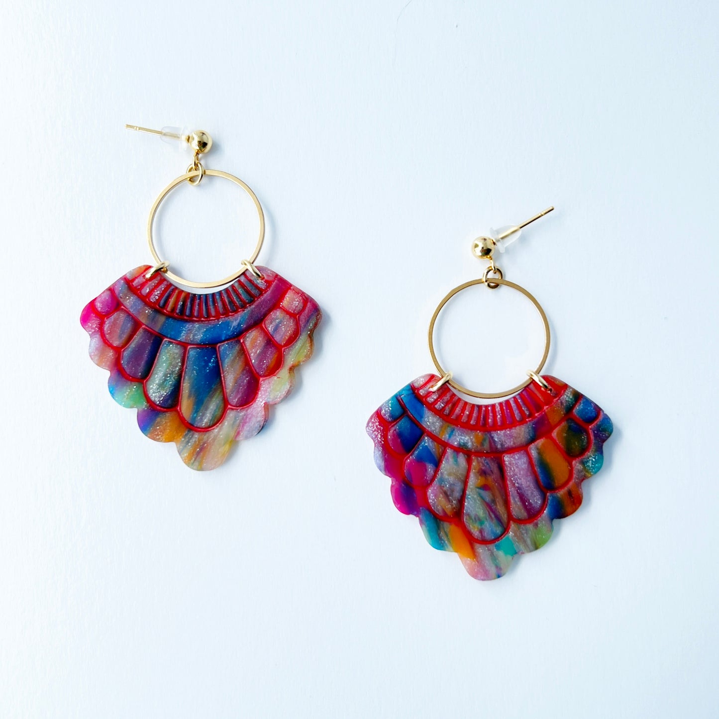 A pair of handmade fan-shaped polymer clay earrings displayed on a white background. The earrings feature gold-plated ball posts and hoops, with semi-transparent rainbow-colored clay and red etched fan-like lines. The marbling effect shows swirls of pink, blue, green, orange, and white.