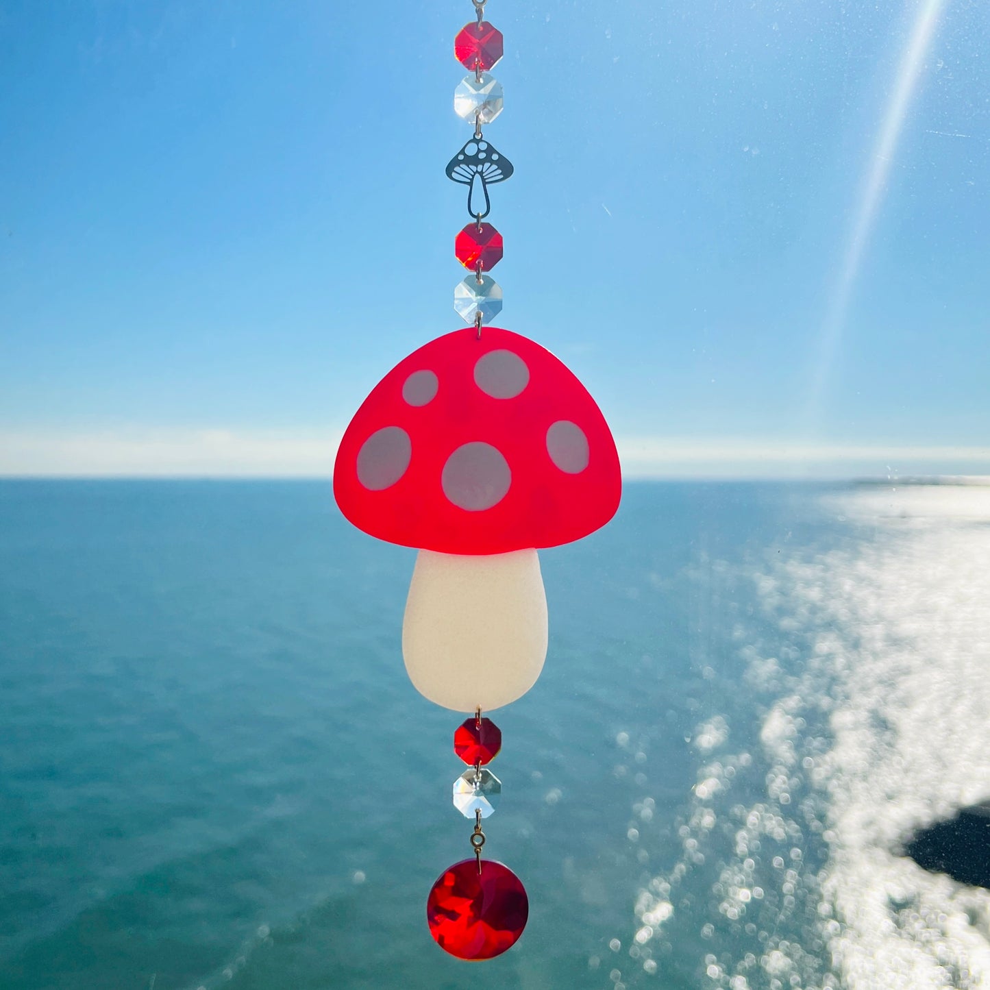 A whimsical handmade mushroom suncatcher crafted from polymer clay, featuring a red and white spotted toadstool design suspended from a gold chain, with a dangling faceted glass crystal that casts rainbows in the sunlight. Ideal for woodland, fairycore, or cottagecore decor.