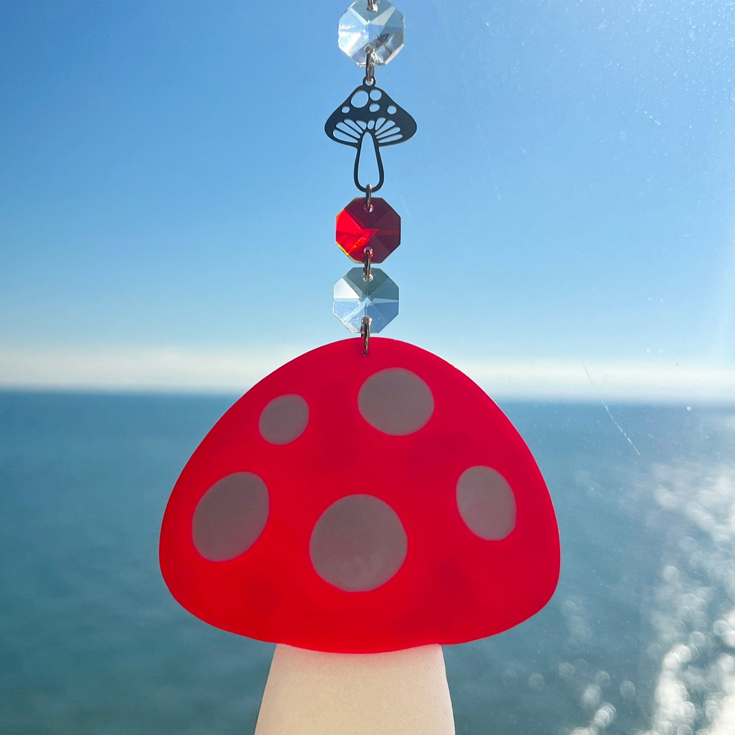 A whimsical handmade mushroom suncatcher crafted from polymer clay, featuring a red and white spotted toadstool design suspended from a gold chain, with a dangling faceted glass crystal that casts rainbows in the sunlight. Ideal for woodland, fairycore, or cottagecore decor.