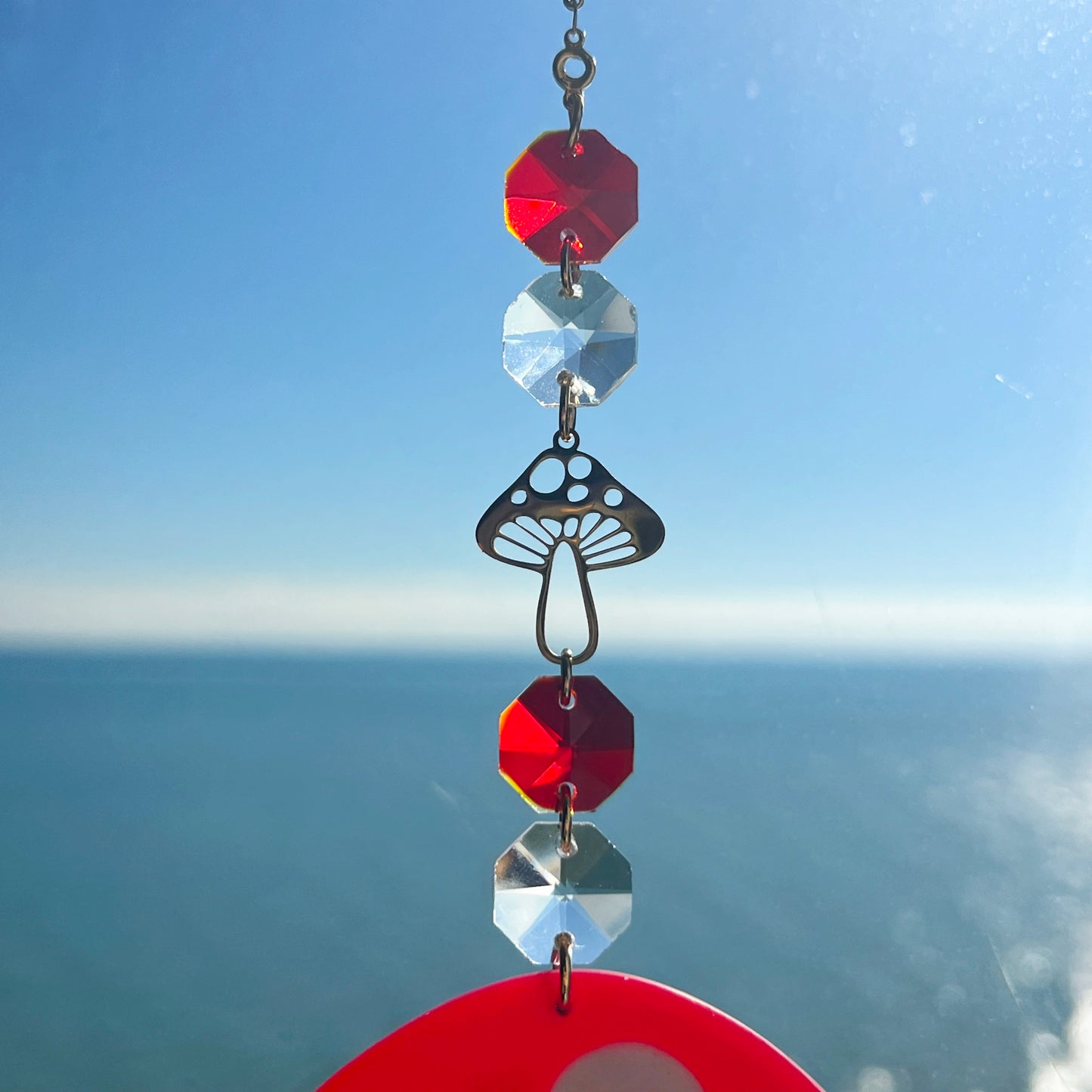 A whimsical handmade mushroom suncatcher crafted from polymer clay, featuring a red and white spotted toadstool design suspended from a gold chain, with a dangling faceted glass crystal that casts rainbows in the sunlight. Ideal for woodland, fairycore, or cottagecore decor.