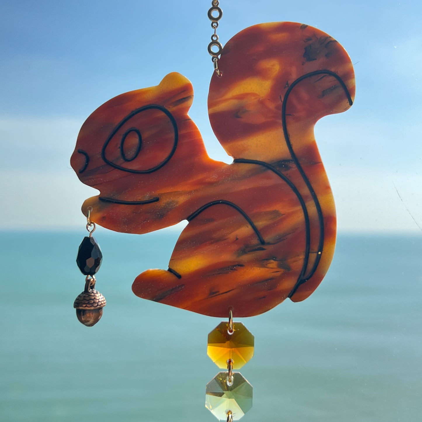 A handmade squirrel suncatcher featuring a brown polymer clay squirrel with a bushy tail and warm autumn tones, hanging from a gold chain with an amber faceted glass maple leaf crystal that reflects rainbow light in the sun. Ideal for woodland, fall, or forest-themed decor.
