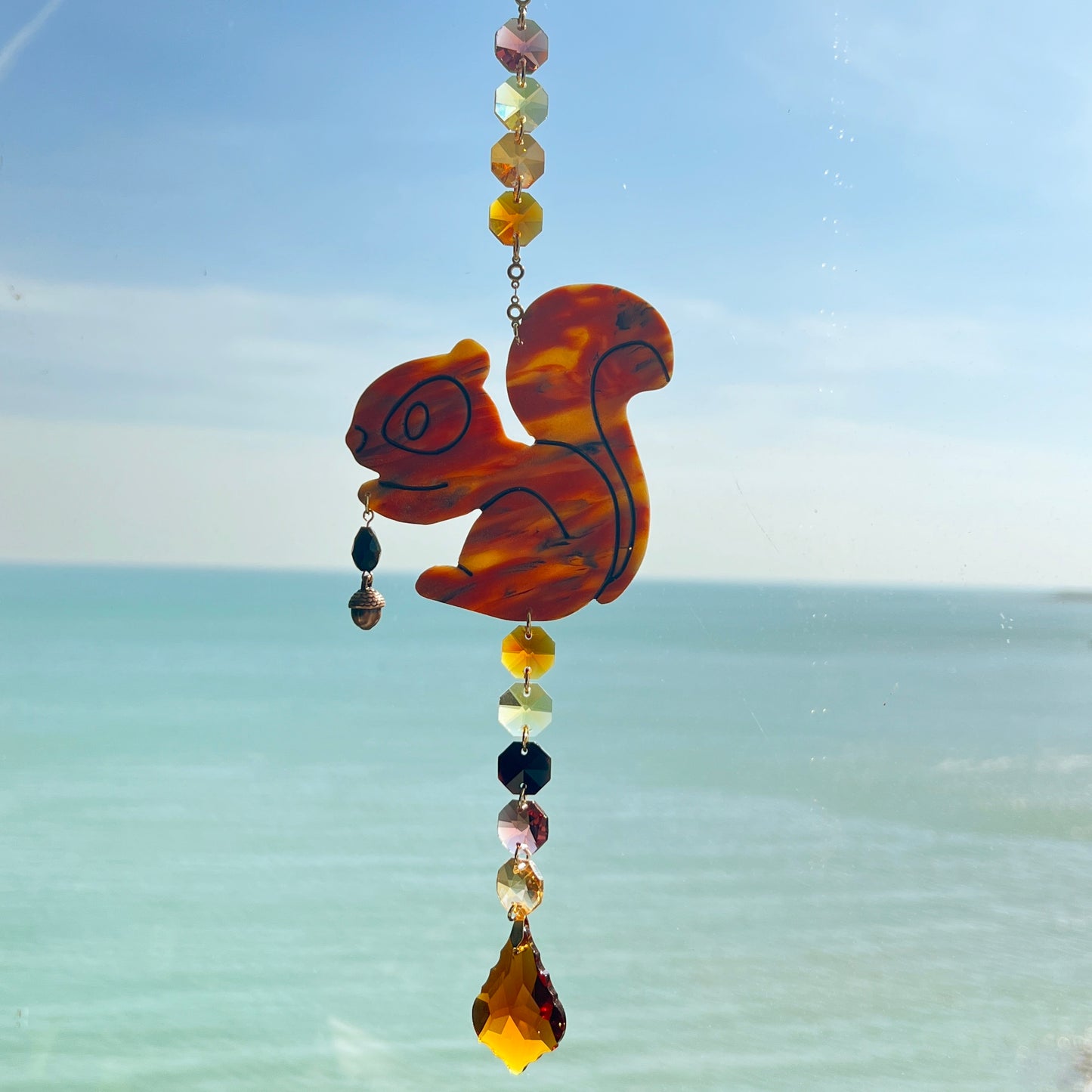 A handmade squirrel suncatcher featuring a brown polymer clay squirrel with a bushy tail and warm autumn tones, hanging from a gold chain with an amber faceted glass maple leaf crystal that reflects rainbow light in the sun. Ideal for woodland, fall, or forest-themed decor.