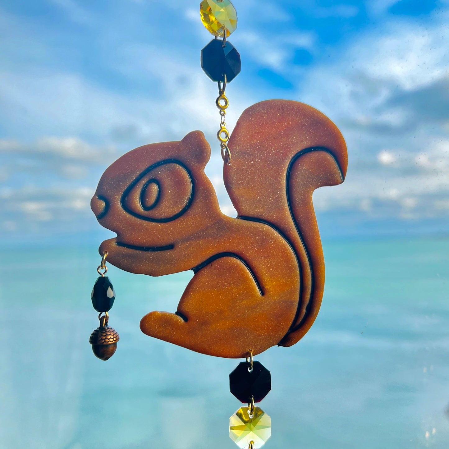 A handmade squirrel suncatcher featuring a brown polymer clay squirrel with a bushy tail and warm autumn tones, hanging from a gold chain with a black faceted glass maple leaf crystal that reflects rainbow light in the sun. Ideal for woodland, fall, or forest-themed decor.