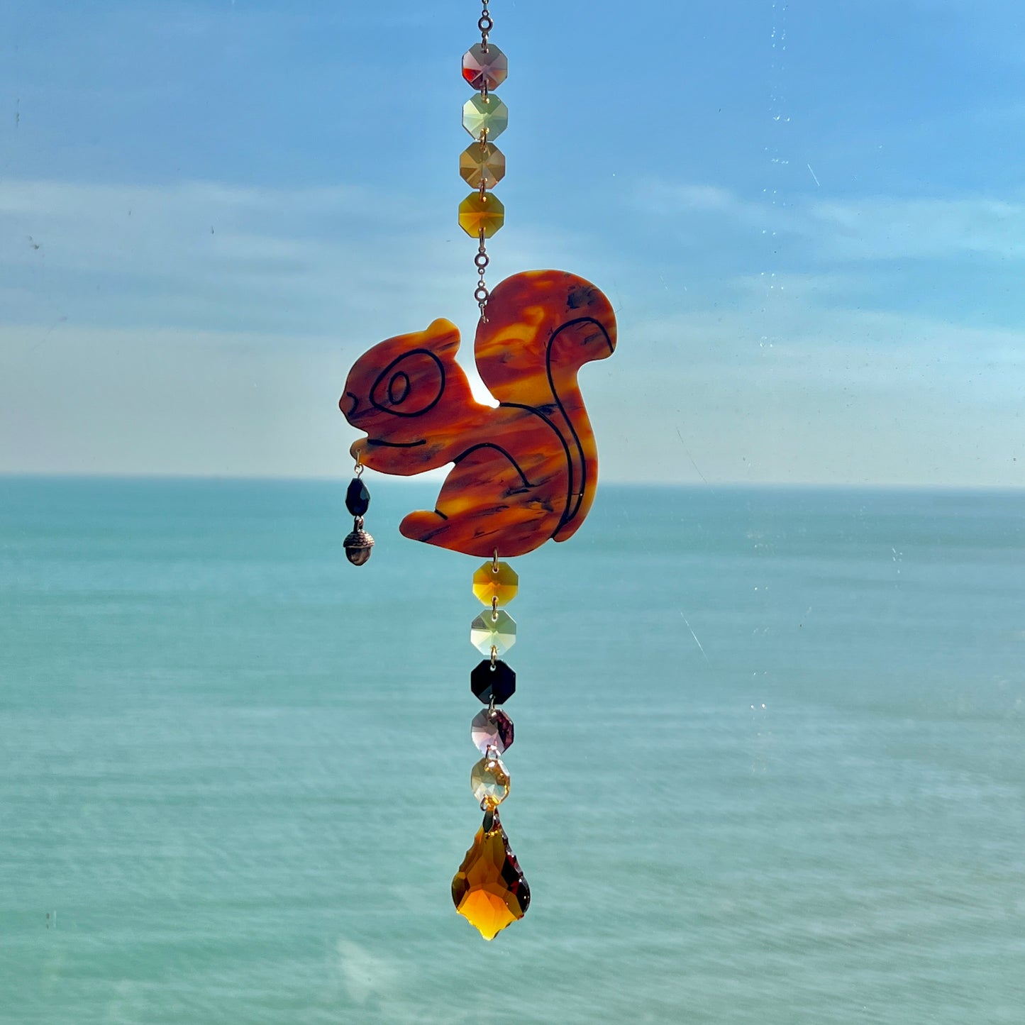 A handmade squirrel suncatcher featuring a brown polymer clay squirrel with a bushy tail and warm autumn tones, hanging from a gold chain with an amber faceted glass maple leaf crystal that reflects rainbow light in the sun. Ideal for woodland, fall, or forest-themed decor.