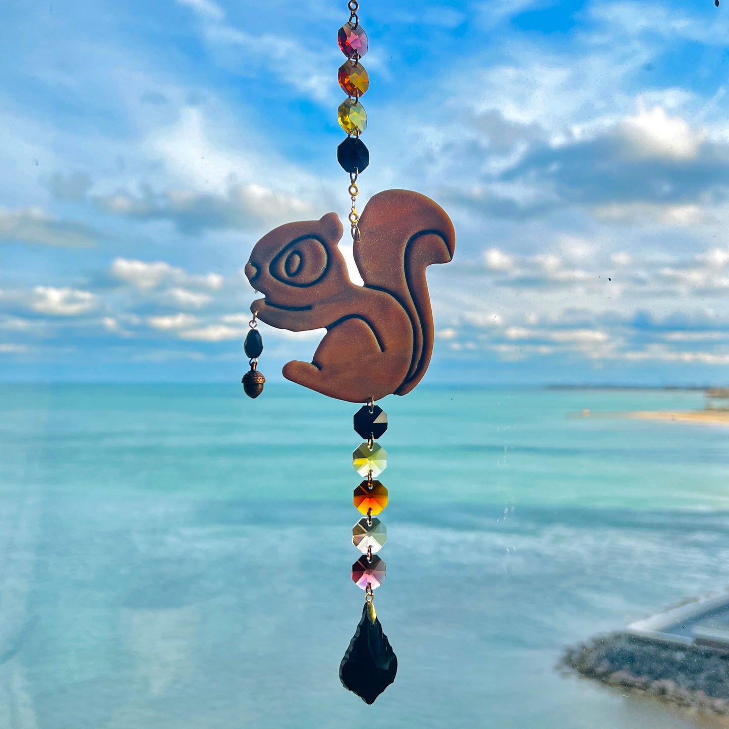 A handmade squirrel suncatcher featuring a brown polymer clay squirrel with a bushy tail and warm autumn tones, hanging from a gold chain with a black faceted glass maple leaf crystal that reflects rainbow light in the sun. Ideal for woodland, fall, or forest-themed decor.