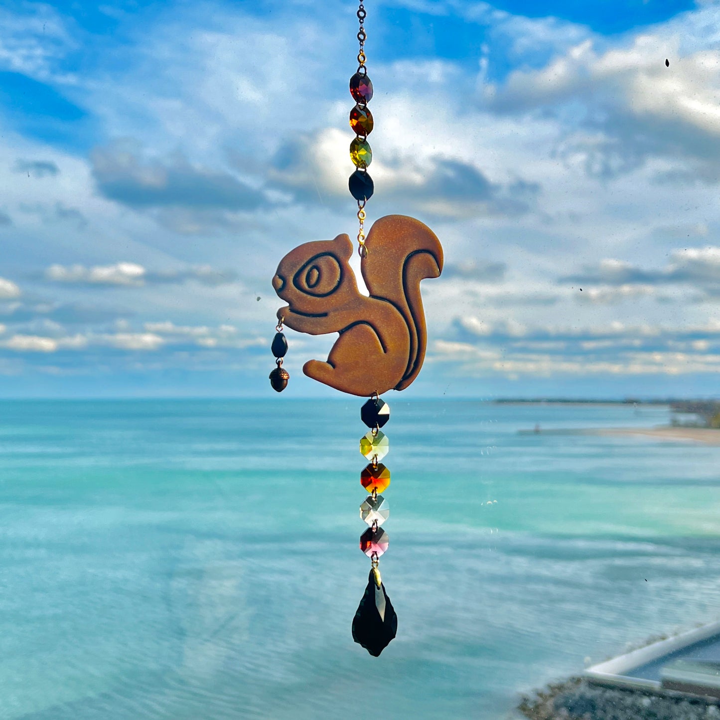 A handmade squirrel suncatcher featuring a brown polymer clay squirrel with a bushy tail and warm autumn tones, hanging from a gold chain with a black faceted glass maple leaf crystal that reflects rainbow light in the sun. Ideal for woodland, fall, or forest-themed decor.