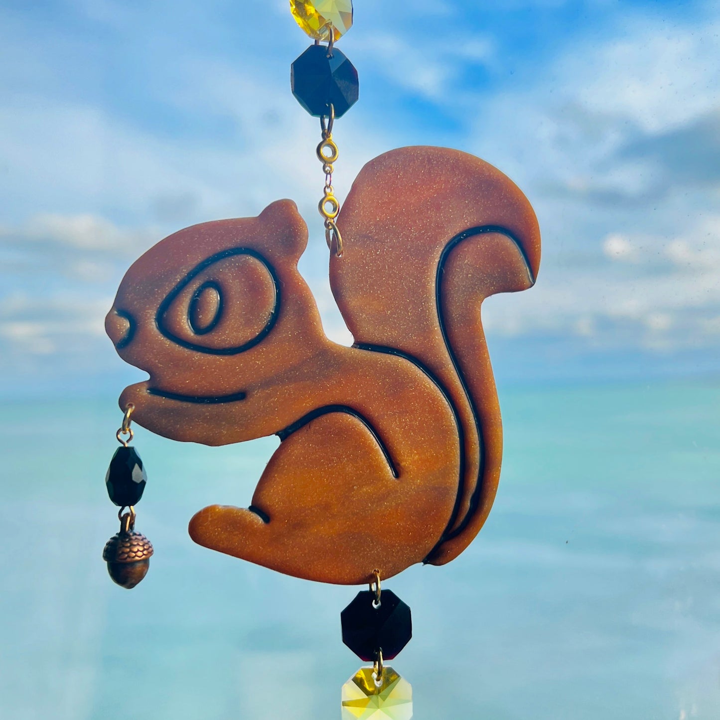 A handmade squirrel suncatcher featuring a brown polymer clay squirrel with a bushy tail and warm autumn tones, hanging from a gold chain with a black faceted glass maple leaf crystal that reflects rainbow light in the sun. Ideal for woodland, fall, or forest-themed decor.
