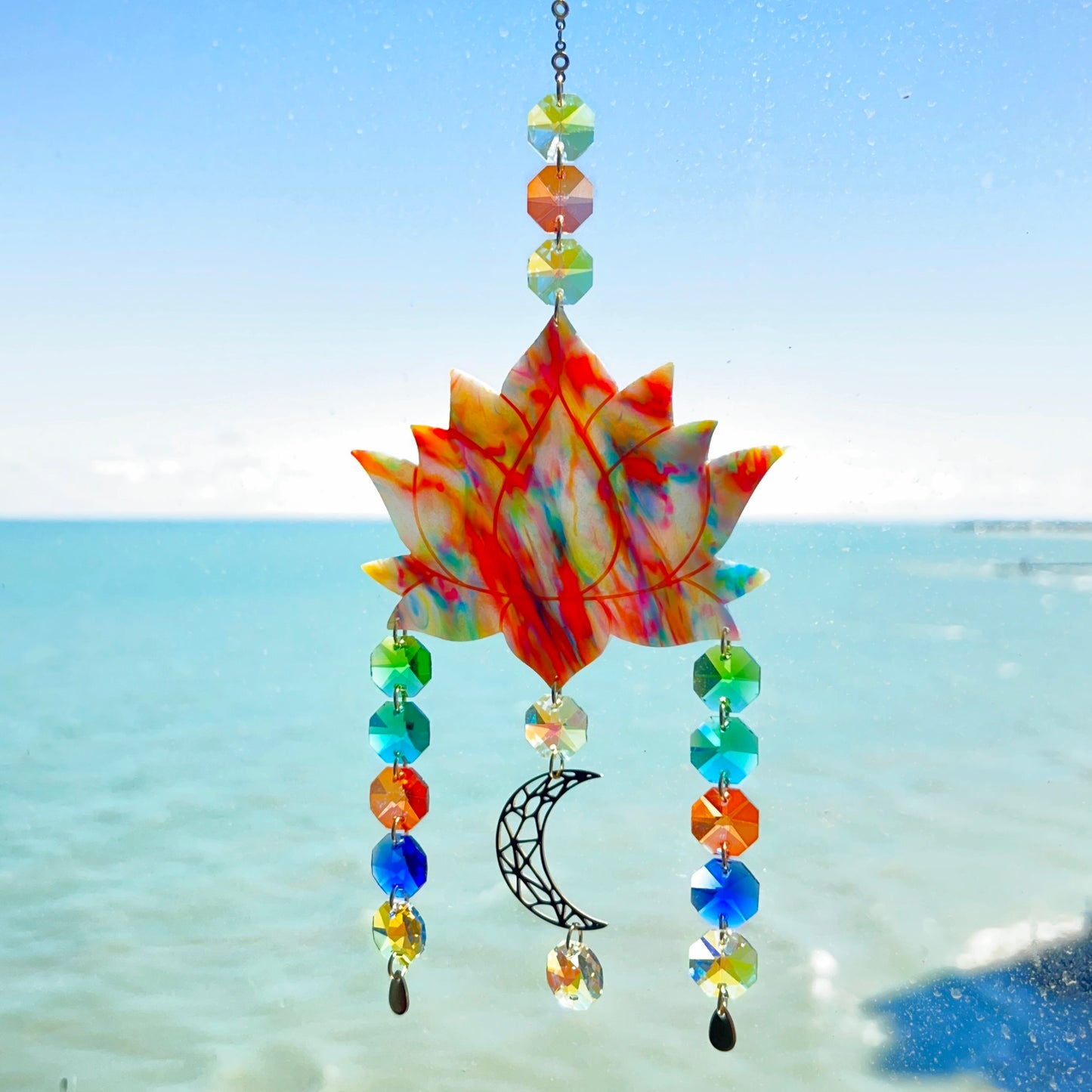 A handmade lotus flower suncatcher crafted from multicolored polymer clay, featuring a stylized lotus bloom suspended from a gold chain with a faceted glass crystal hanging below, designed to reflect rainbow light in the sun. Ideal for meditation rooms, spiritual decor, or peaceful window displays.
