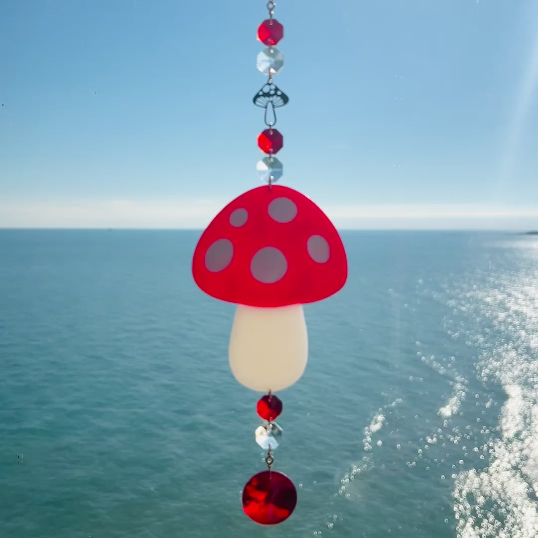 Video showing a whimsical handmade mushroom suncatcher crafted from polymer clay, featuring a red and white spotted toadstool design suspended from a gold chain, with a dangling faceted glass crystal that casts rainbows in the sunlight. Ideal for woodland, fairycore, or cottagecore decor.