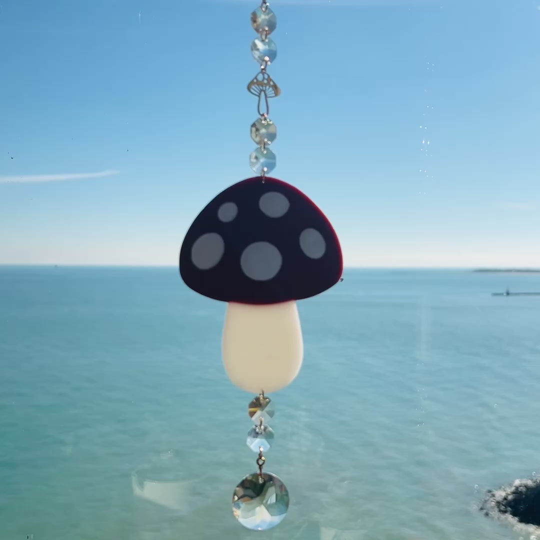 Video of a whimsical handmade mushroom suncatcher crafted from polymer clay, featuring a brown and white spotted toadstool design suspended from a gold chain, with a dangling faceted glass crystal that casts rainbows in the sunlight. Ideal for woodland, fairycore, or cottagecore decor.