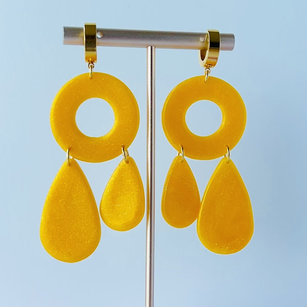 Video of a pair of handmade dangle earrings  hanging on a gold T-bar spinning slowly in a circle to show all side.  The earrings feature semi-transparent yellow polymer clay with a subtle shimmer. Each earring has a round donut-shaped top connected to two teardrop shapes of different sizes. They are attached to chunky, gold-plated stainless steel leverback hoops