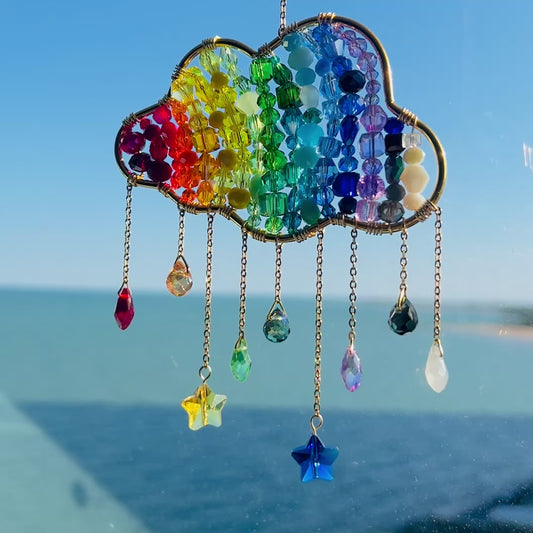 Video of a handmade rainbow suncatcher shaped like a cloud, crafted with a wire frame filled with rows of multicolored glass beads in red, orange, yellow, green, blue, and violet. Dangling from the bottom are gold chains with various glass bead charms, including teardrops, stars, and faceted gems in coordinating rainbow colors. The suncatcher is shown hanging in a sunlit window with a bright blue sky and water in the background.