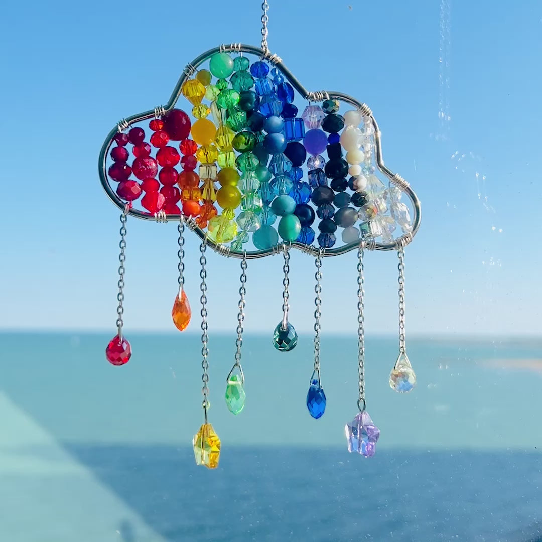 Video of a A handmade rainbow suncatcher shaped like a cloud, crafted with a silver wire frame filled with rows of multicolored glass beads in red, orange, yellow, green, blue, and violet. Dangling from the bottom are silver chains with various glass bead charms, including teardrops, stars, and faceted gems in coordinating rainbow colors. The suncatcher is shown hanging in a sunlit window with a bright blue sky and water in the background.