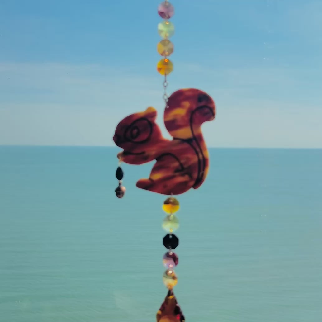 Video of a handmade squirrel suncatcher featuring a brown polymer clay squirrel with a bushy tail and warm autumn tones, hanging from a gold chain with an amber faceted glass maple leaf crystal that reflects rainbow light in the sun. Ideal for woodland, fall, or forest-themed decor.