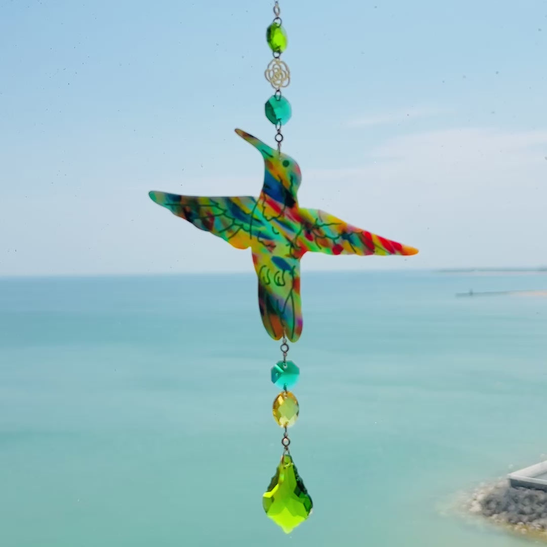 Video of a handmade hummingbird suncatcher crafted from colorful polymer clay, featuring a detailed hummingbird design in shades of green, blue, and pink, suspended from a gold chain with a faceted green maple leaf glass crystal that casts rainbows in the sunlight. Perfect for nature lovers and whimsical window decor.