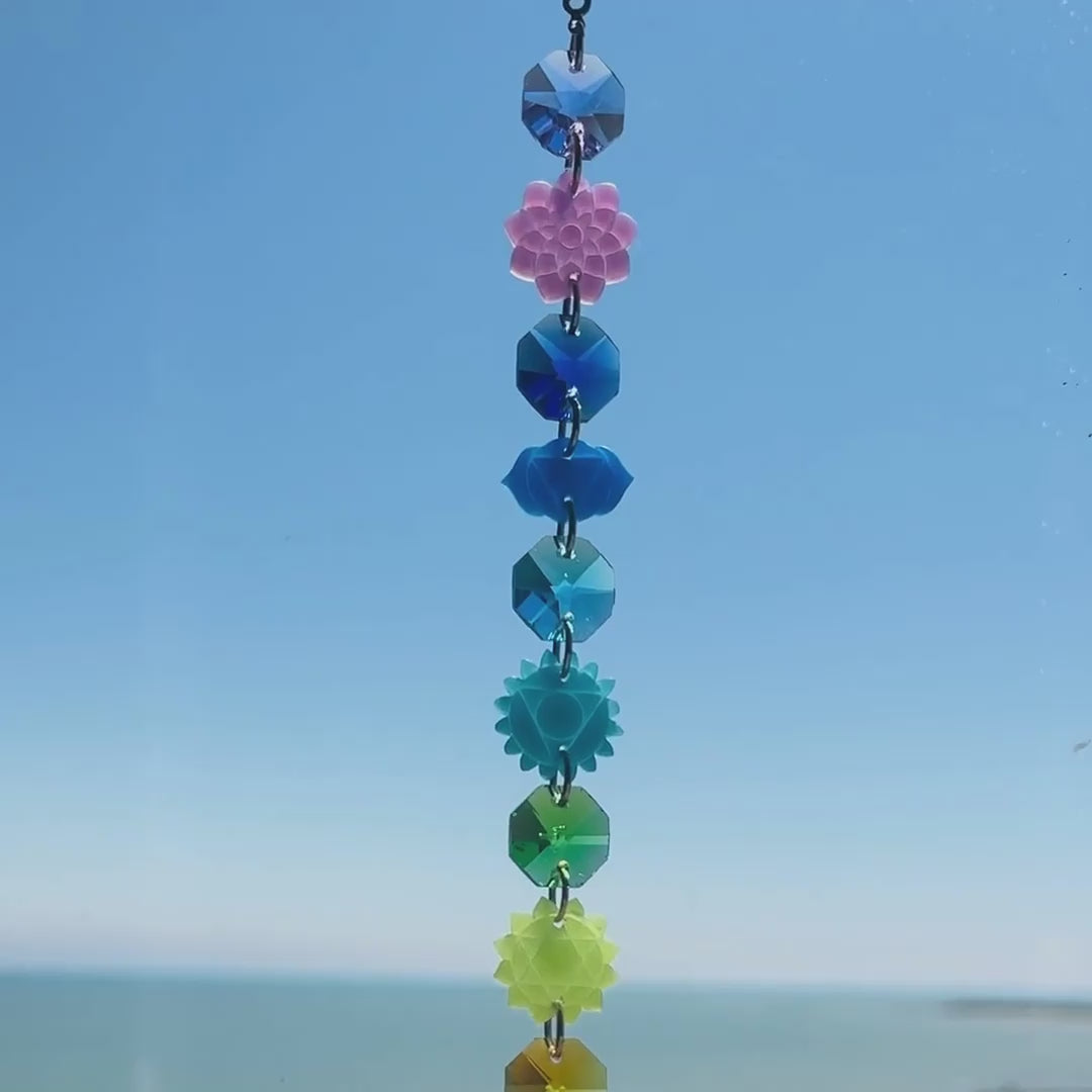 Video of a handmade chakra suncatcher featuring seven stacked polymer clay beads in the colors of the chakras, strung vertically and finished with a hanging faceted glass crystal that reflects rainbows when hit by sunlight. Designed for spiritual, boho, or meditative decor spaces.