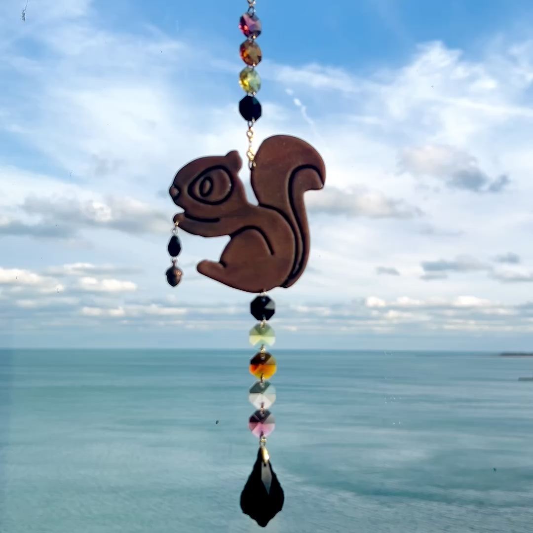 Video of a handmade squirrel suncatcher featuring a brown polymer clay squirrel with a bushy tail and warm autumn tones, hanging from a gold chain with a black faceted glass maple leaf crystal that reflects rainbow light in the sun. Ideal for woodland, fall, or forest-themed decor.