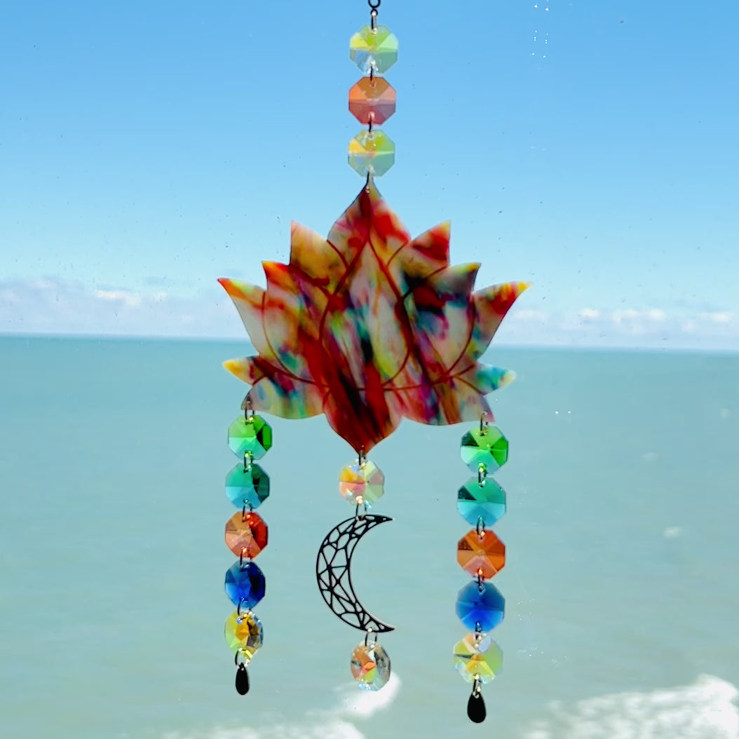 Video of a handmade lotus flower suncatcher crafted from multicolored polymer clay, featuring a stylized lotus bloom suspended from a gold chain with a faceted glass crystal hanging below, designed to reflect rainbow light in the sun. Ideal for meditation rooms, spiritual decor, or peaceful window displays.
